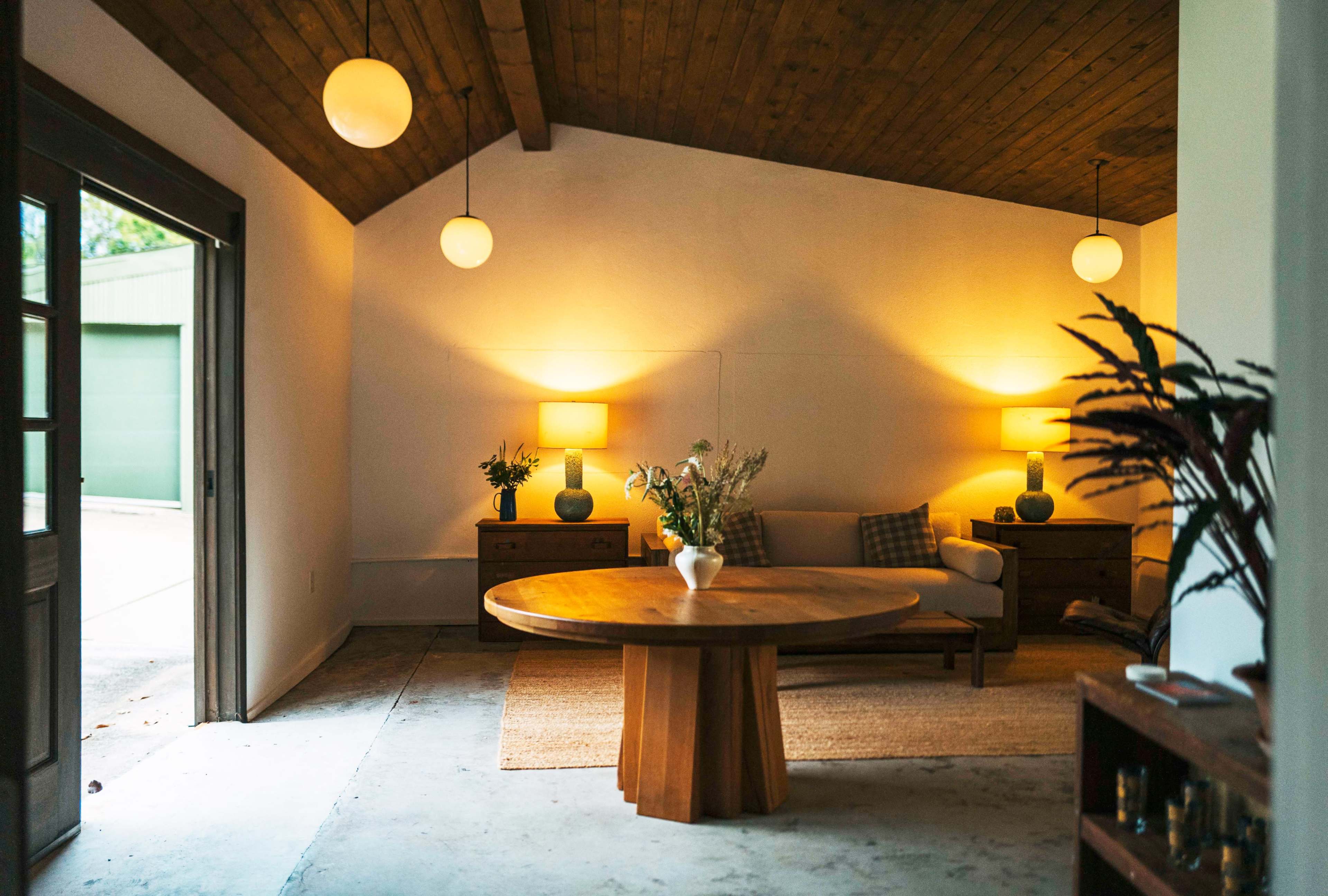 The image shows a minimalist interior space with a round wooden table at the center, flanked by soft lighting from two lamps and a cozy seating area.