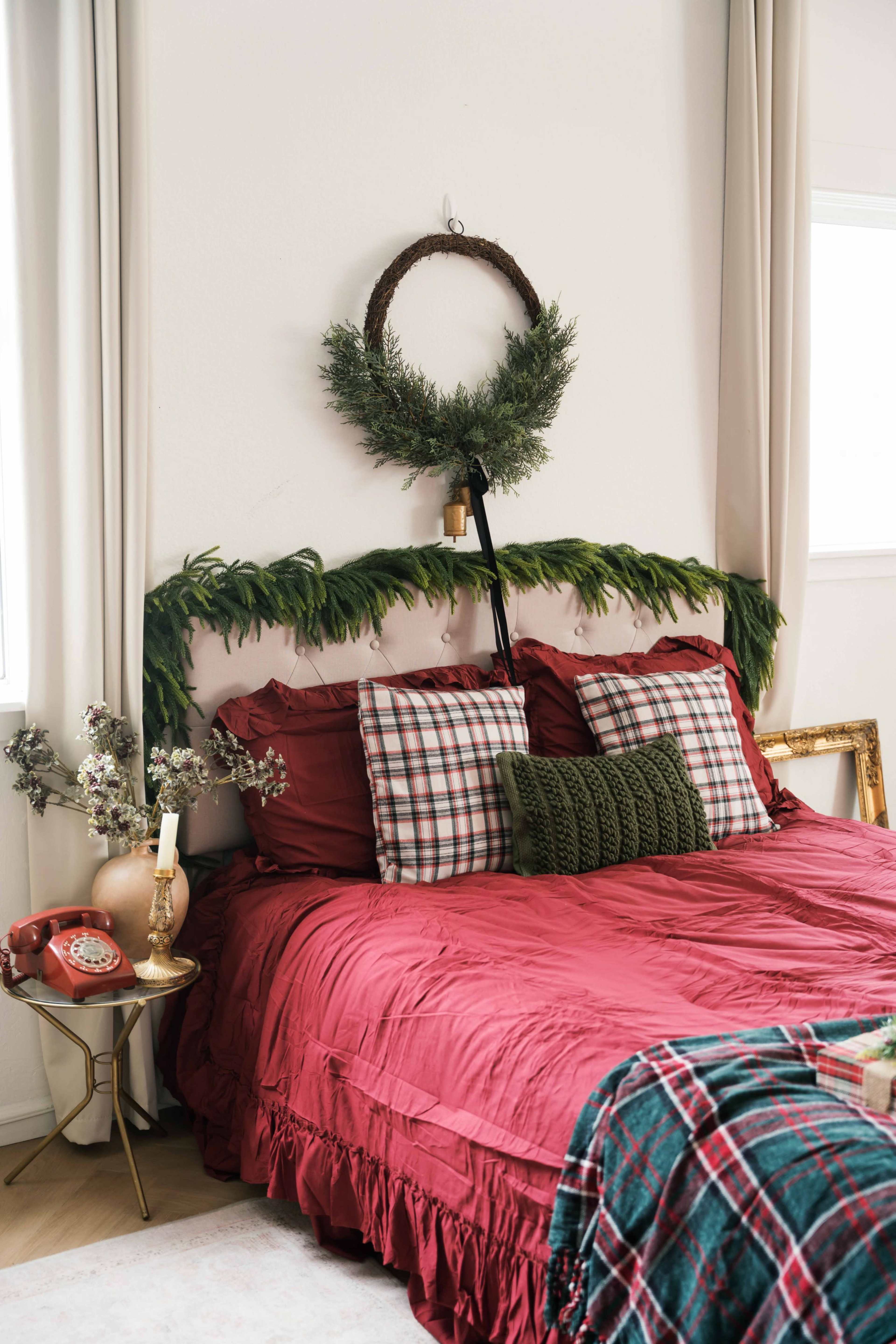 A cozy bedroom features a red bedspread with patterned pillows, a green wreath hanging above the headboard, and decorative elements like a vintage phone and a small vase of flowers on a bedside table.