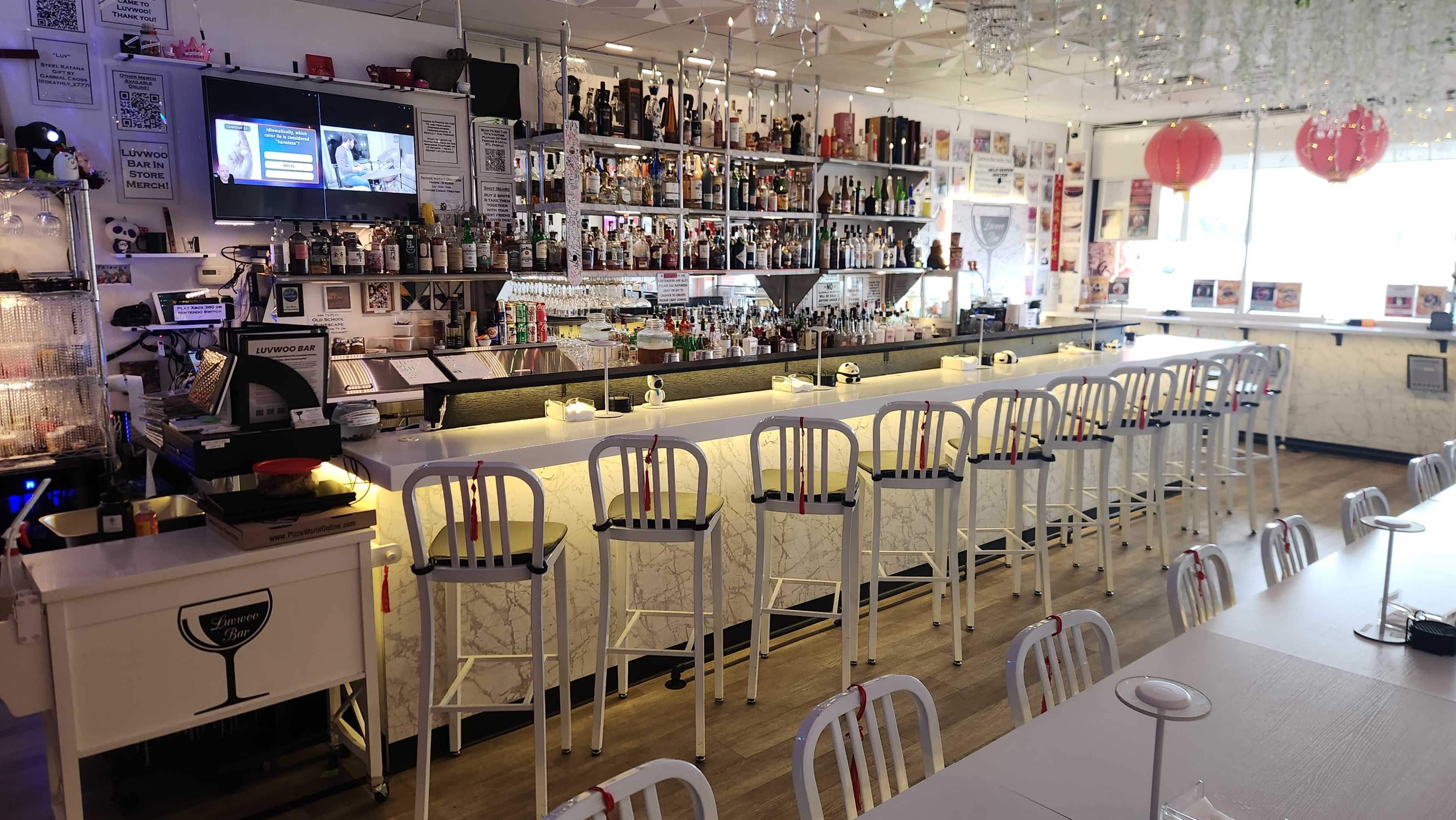 The image shows a modern bar area with a long white marble counter, high stools, and shelves stocked with various bottles and drinks.