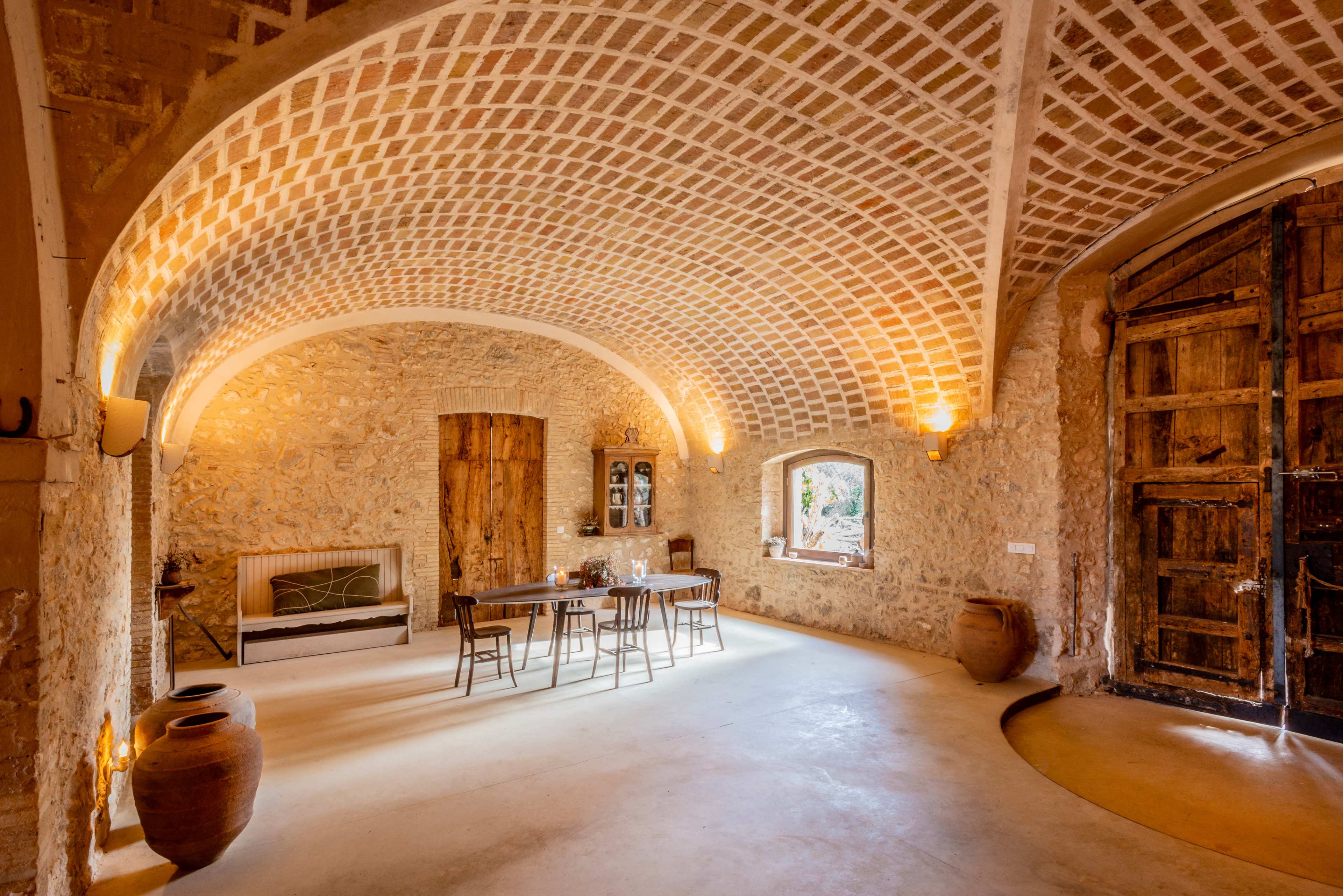 The interior of a rustic stone building features a vaulted ceiling, a long dining table, and wooden doors, with warm lighting illuminating the space.