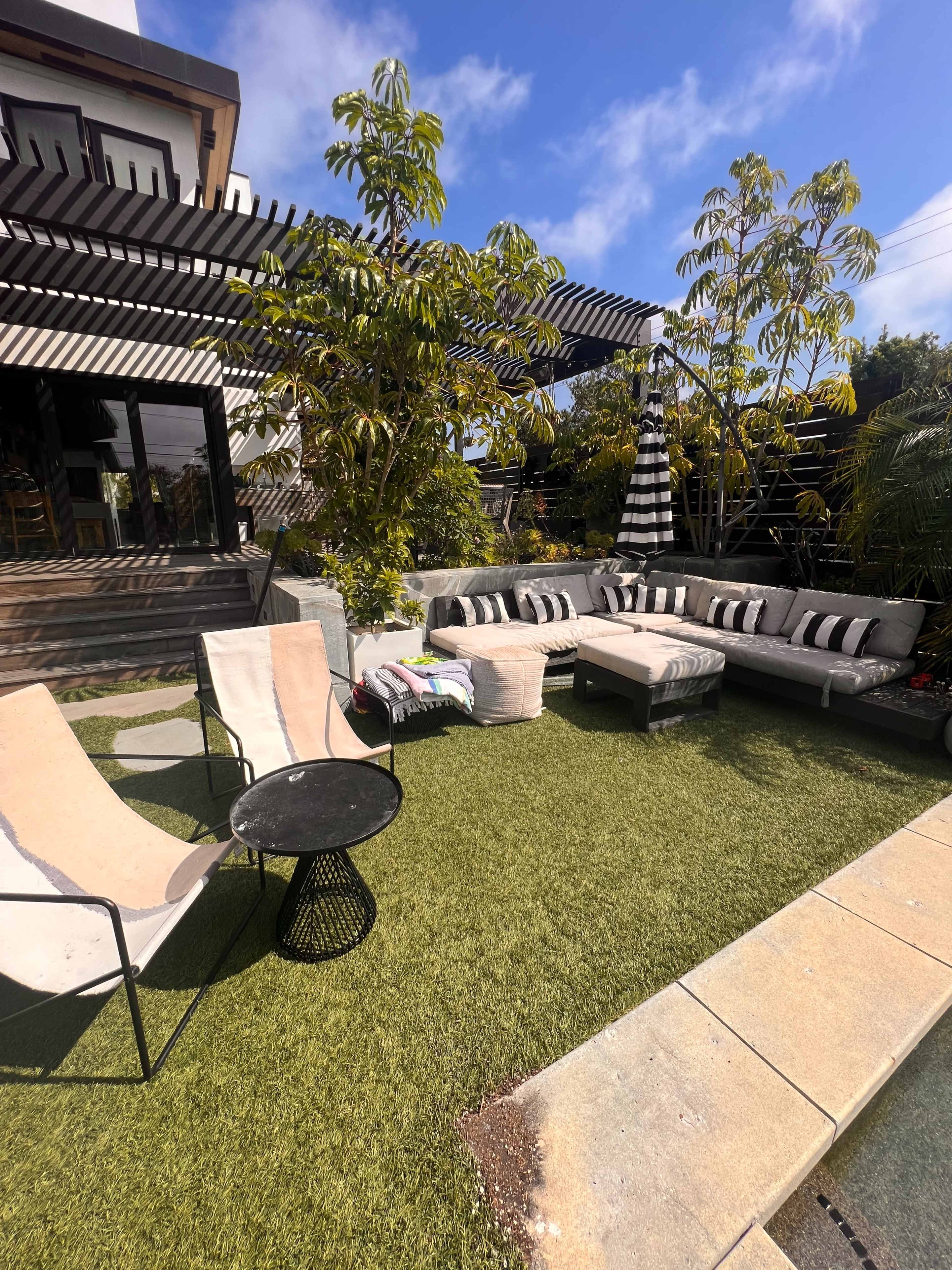 A landscaped outdoor seating area featuring a cozy arrangement of sofas, chairs, and tables surrounded by greenery and a partly cloudy sky.