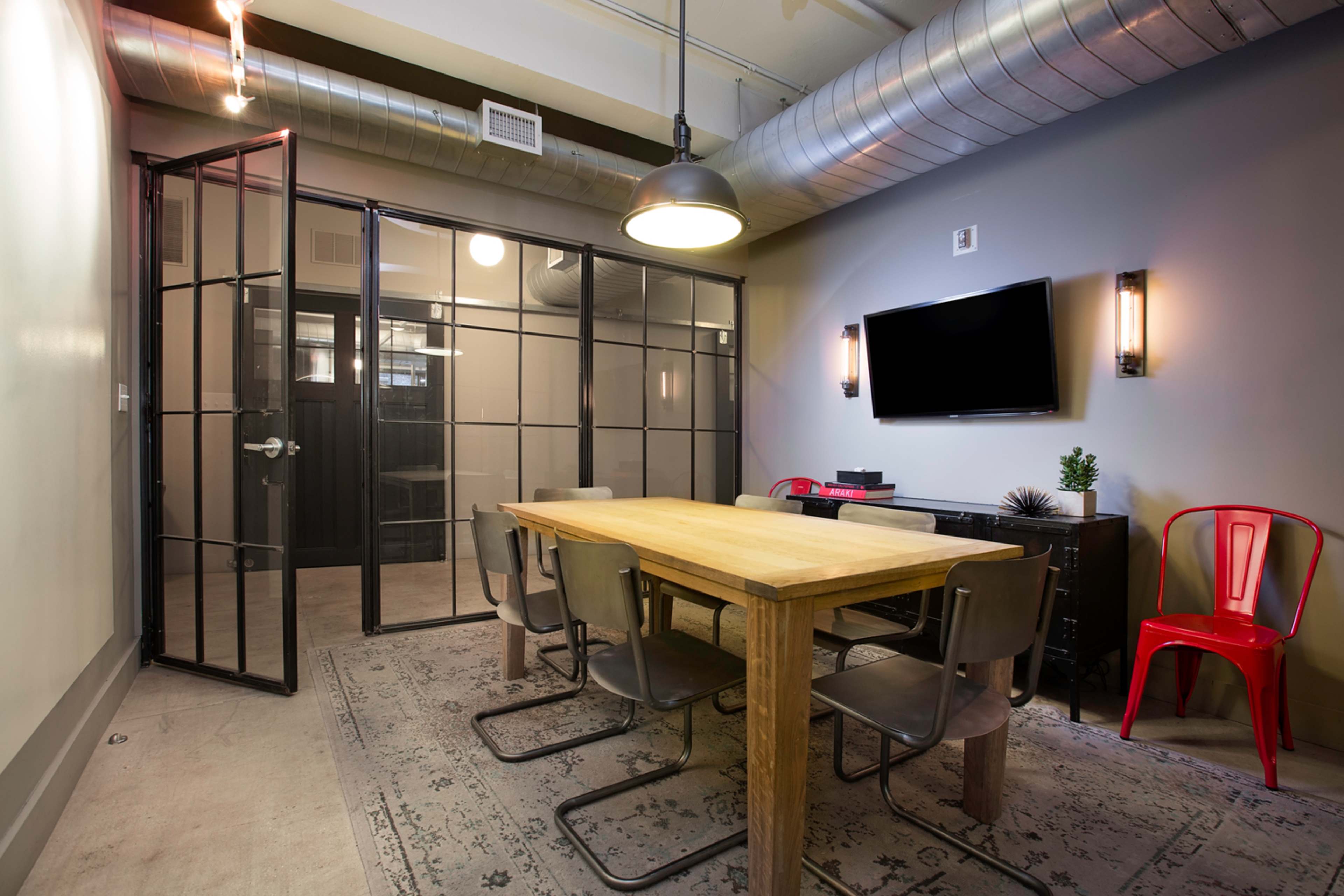A conference room features a wooden table surrounded by metal chairs, with glass-paneled walls and a wall-mounted television.