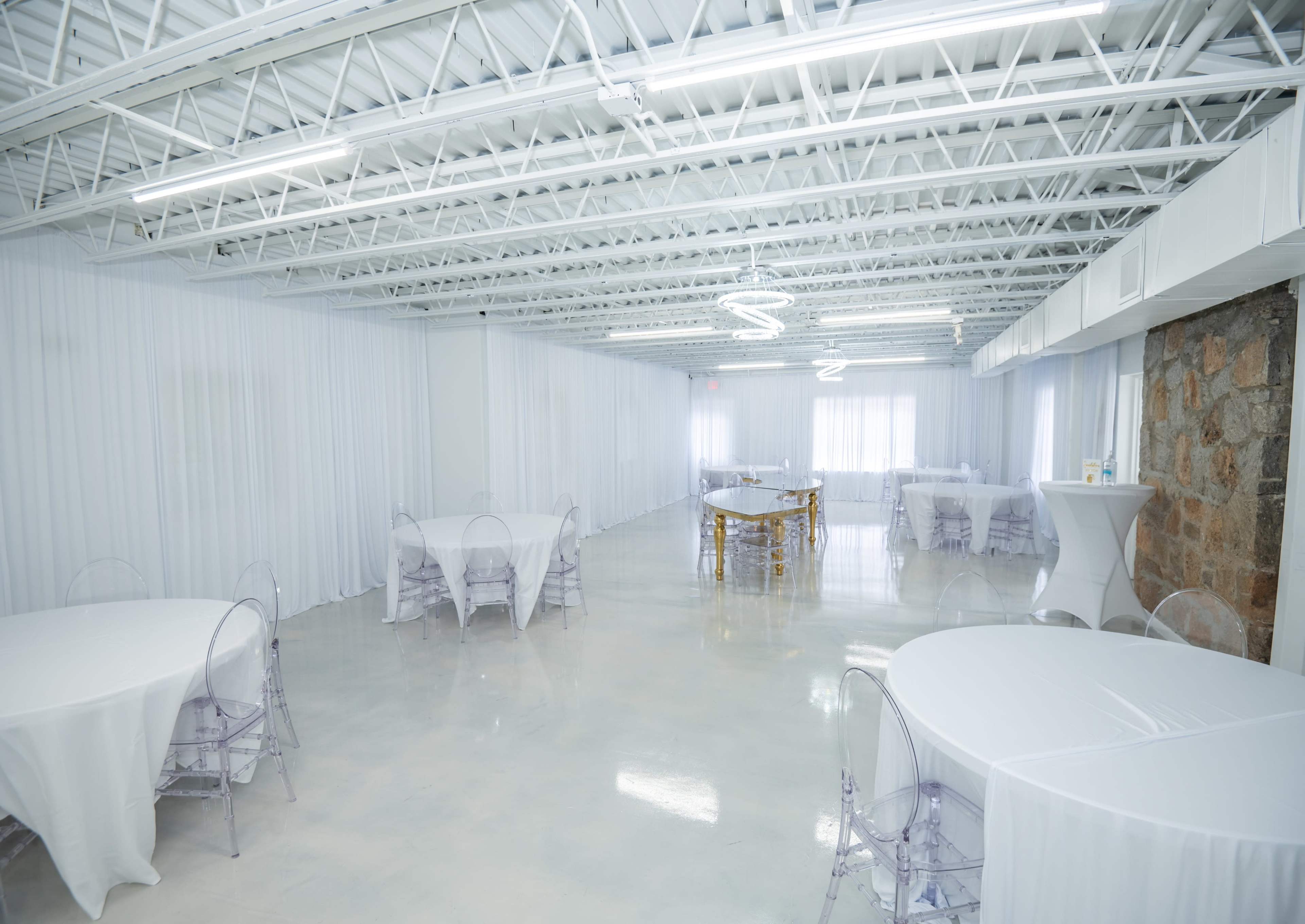 The image shows a spacious, well-lit event venue featuring several round tables with white tablecloths and transparent chairs.