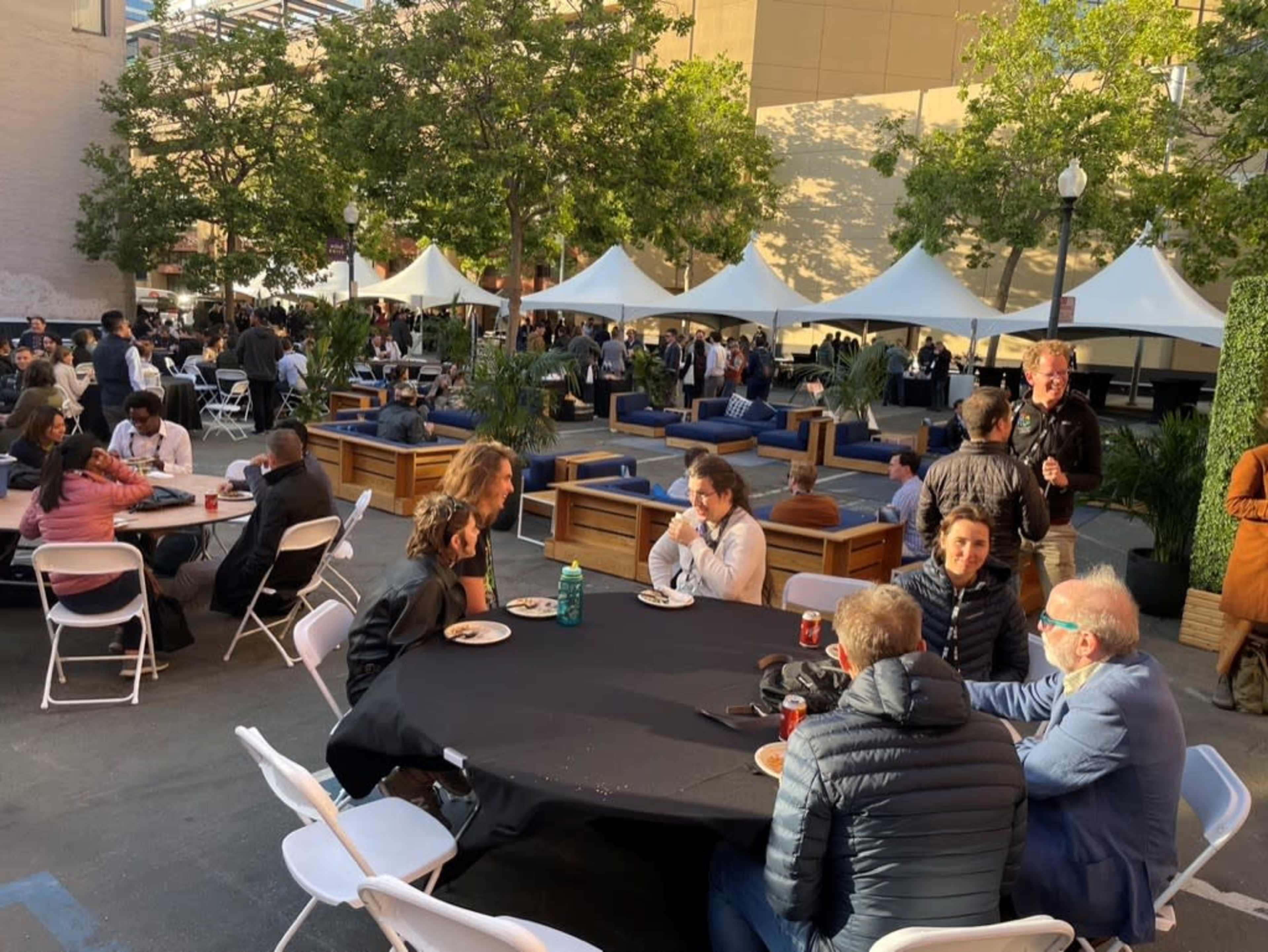 A gathering of people is seated at tables in an outdoor space with tents and seating areas in the background.