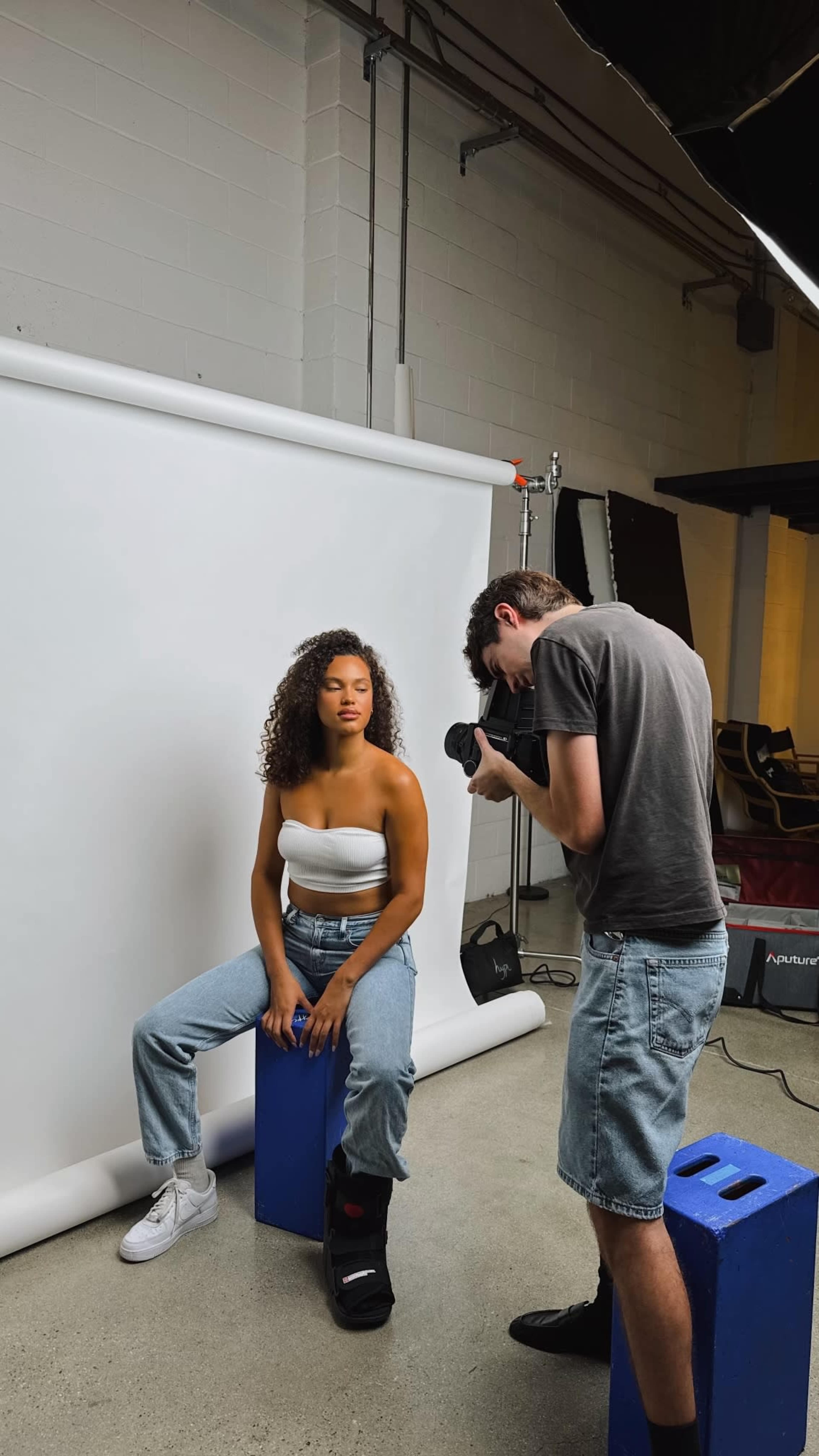 A photographer captures a model sitting on a blue block against a white backdrop in a studio setting.