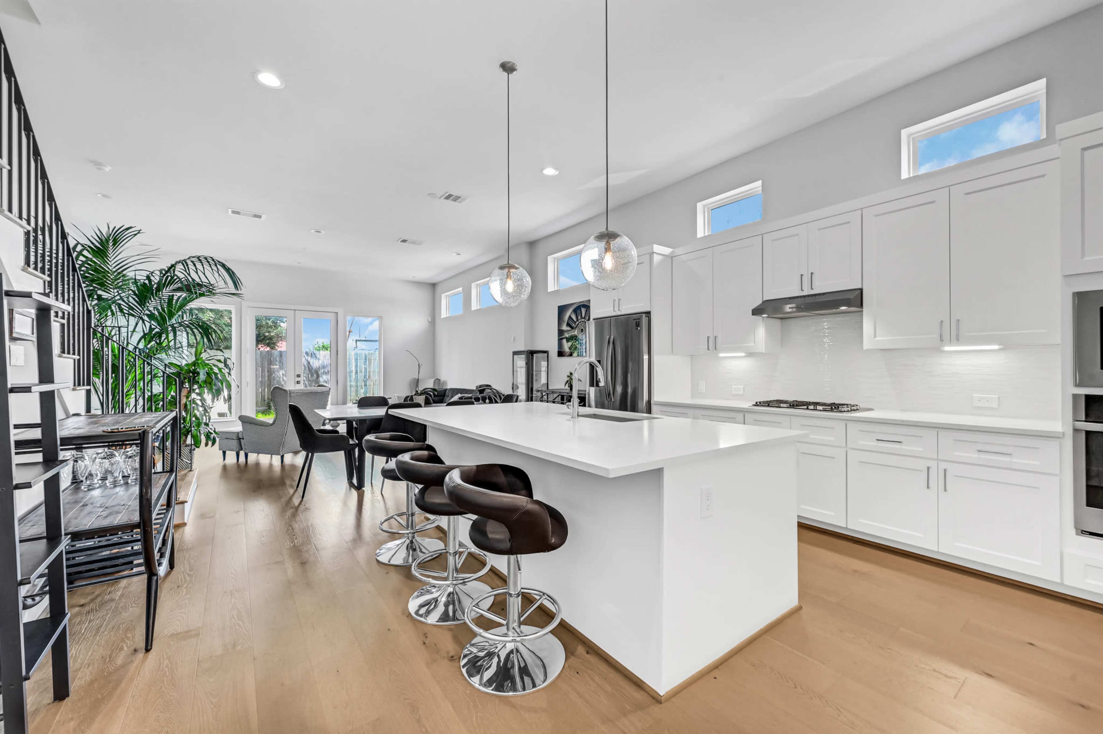 The image shows a modern kitchen and dining area with a large island featuring bar stools, white cabinetry, large windows, and plants.