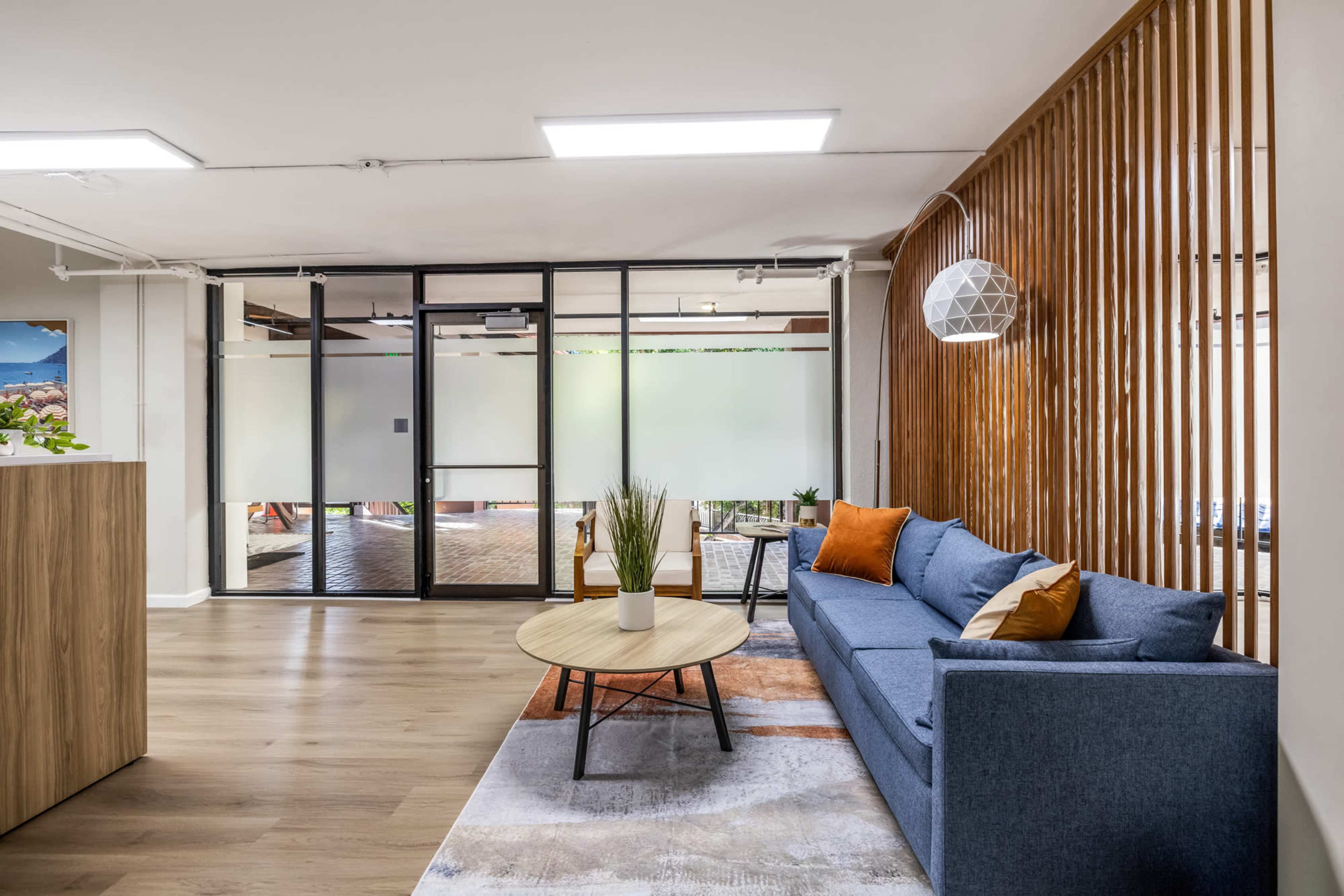 A modern office interior featuring a blue sofa, a round coffee table, and a wooden screen, with glass doors leading to a brighter area.
