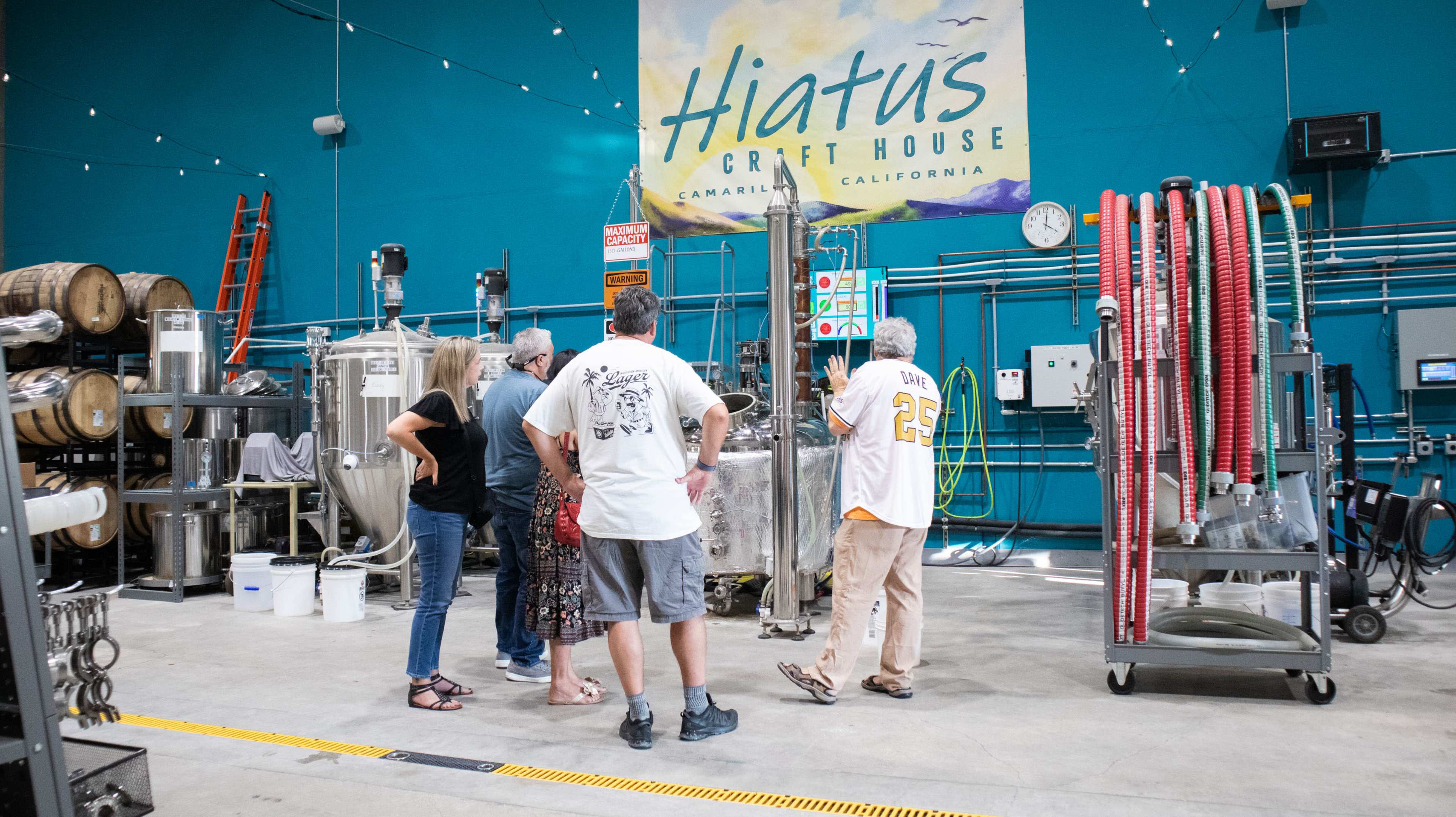 A group of five people is gathered around brewing equipment in a bright blue craft brewery, with a large sign reading "Hiatus Craft House" above them.