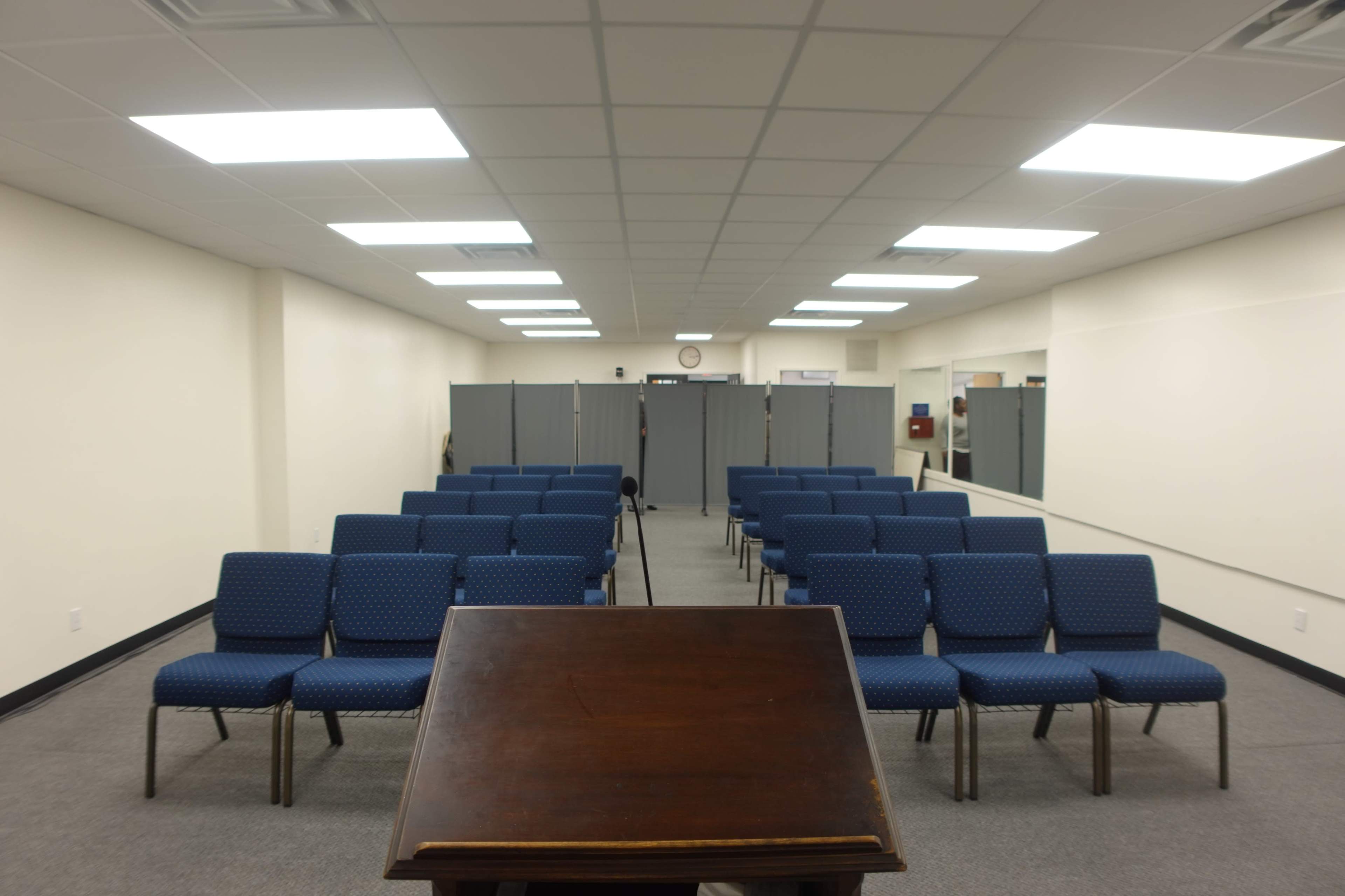 A room with rows of blue chairs arranged in front of a wooden lectern, with partitions and a clock visible in the background.