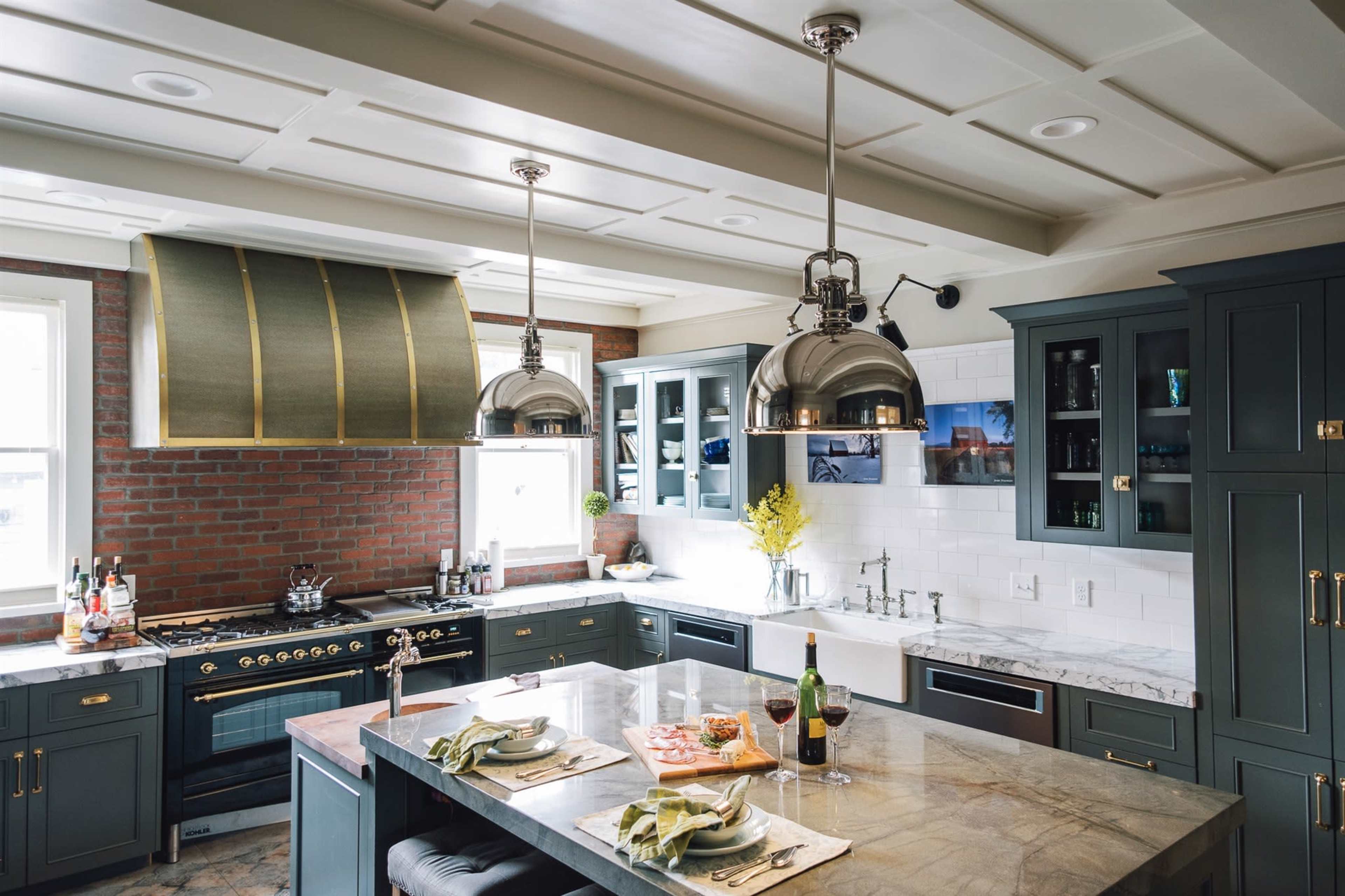 The kitchen features a central island with a marble countertop, stainless steel appliances, and dark cabinetry, complemented by a large hood and brick accent wall.