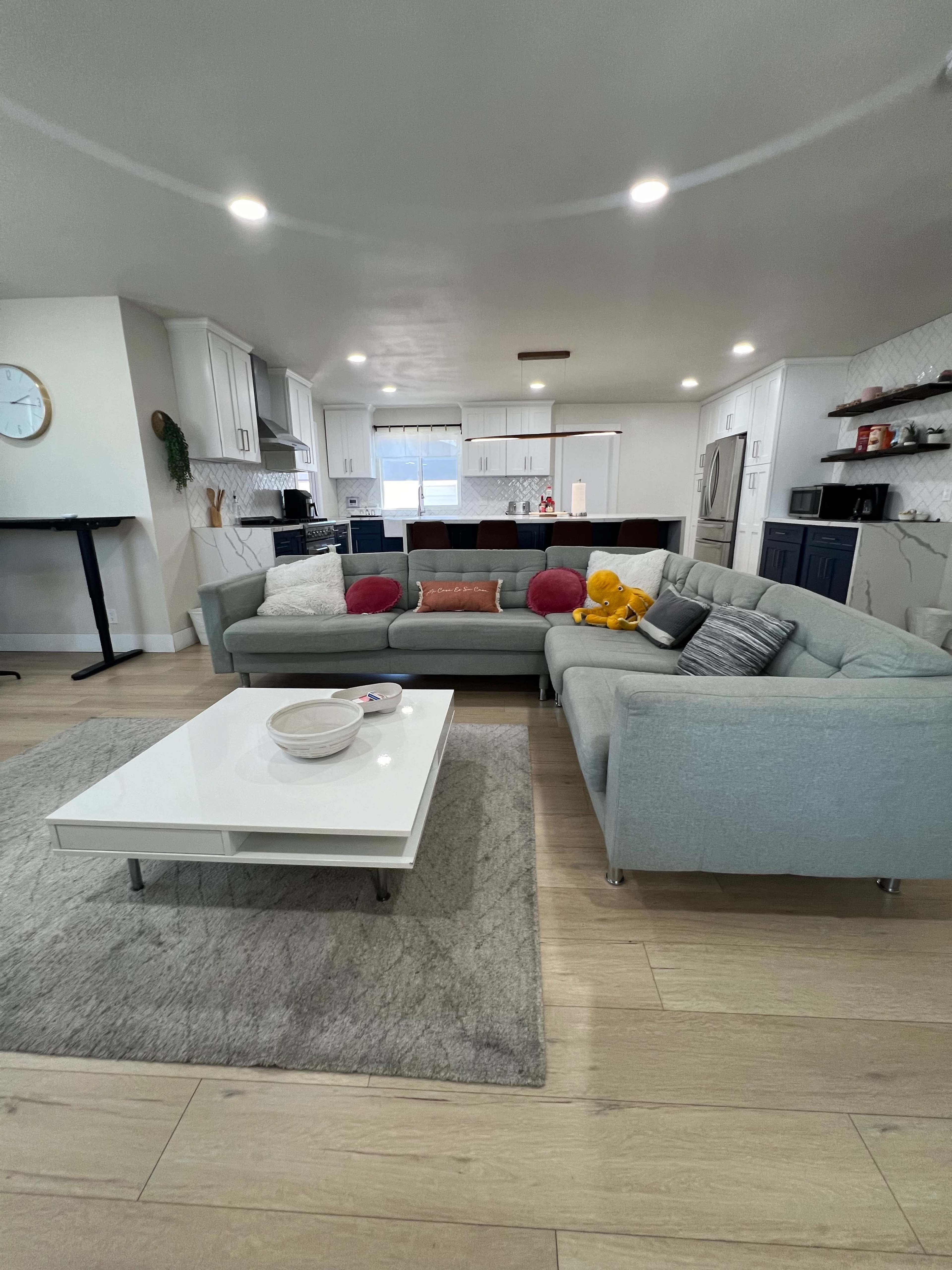 A modern living room features a light gray sectional sofa with colorful pillows, a white coffee table on a gray rug, and a view of a kitchen with white cabinets in the background.