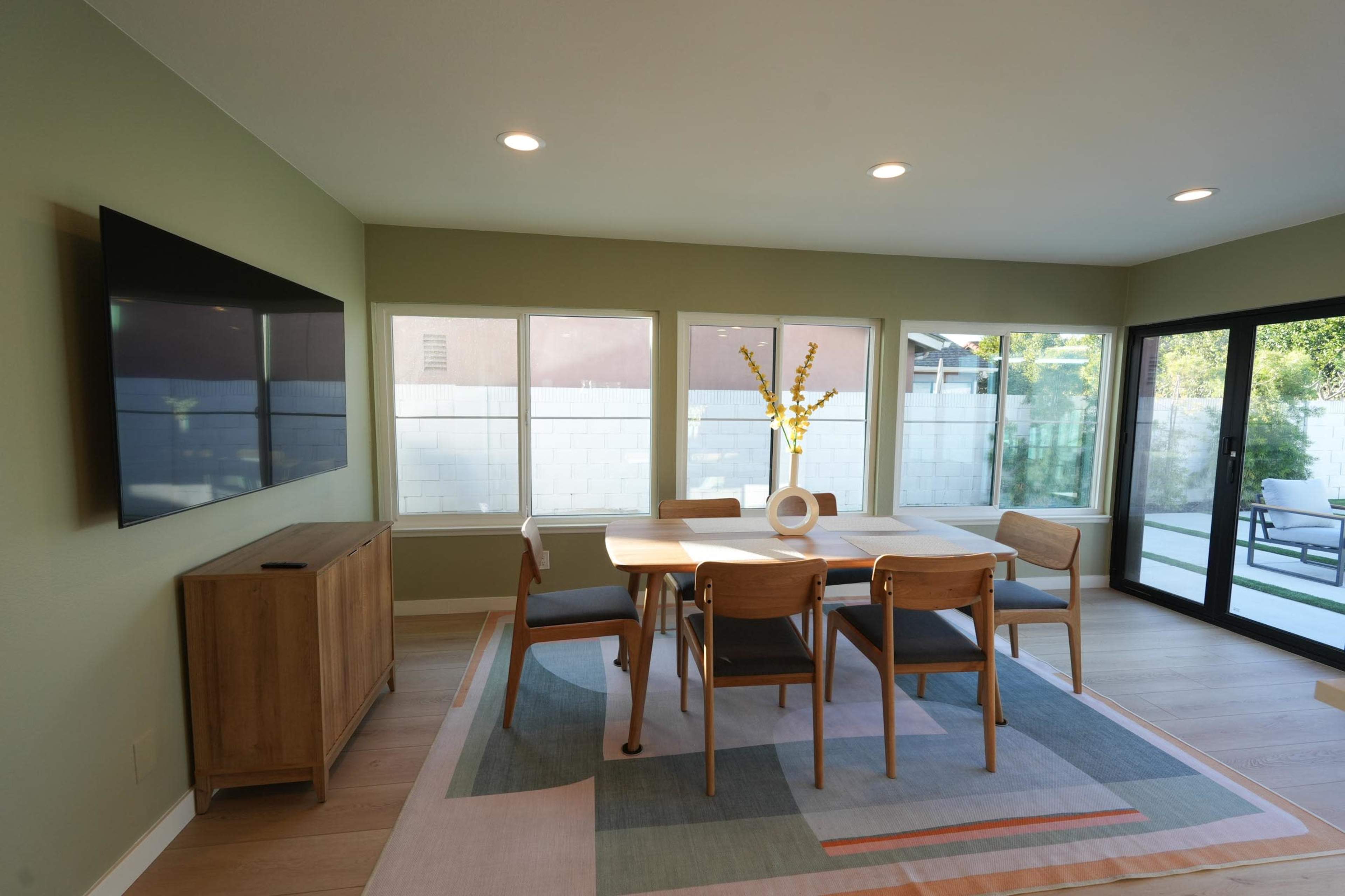 The room features a dining table with four chairs, a sideboard, and a large TV mounted on the wall, with windows and sliding doors providing natural light.