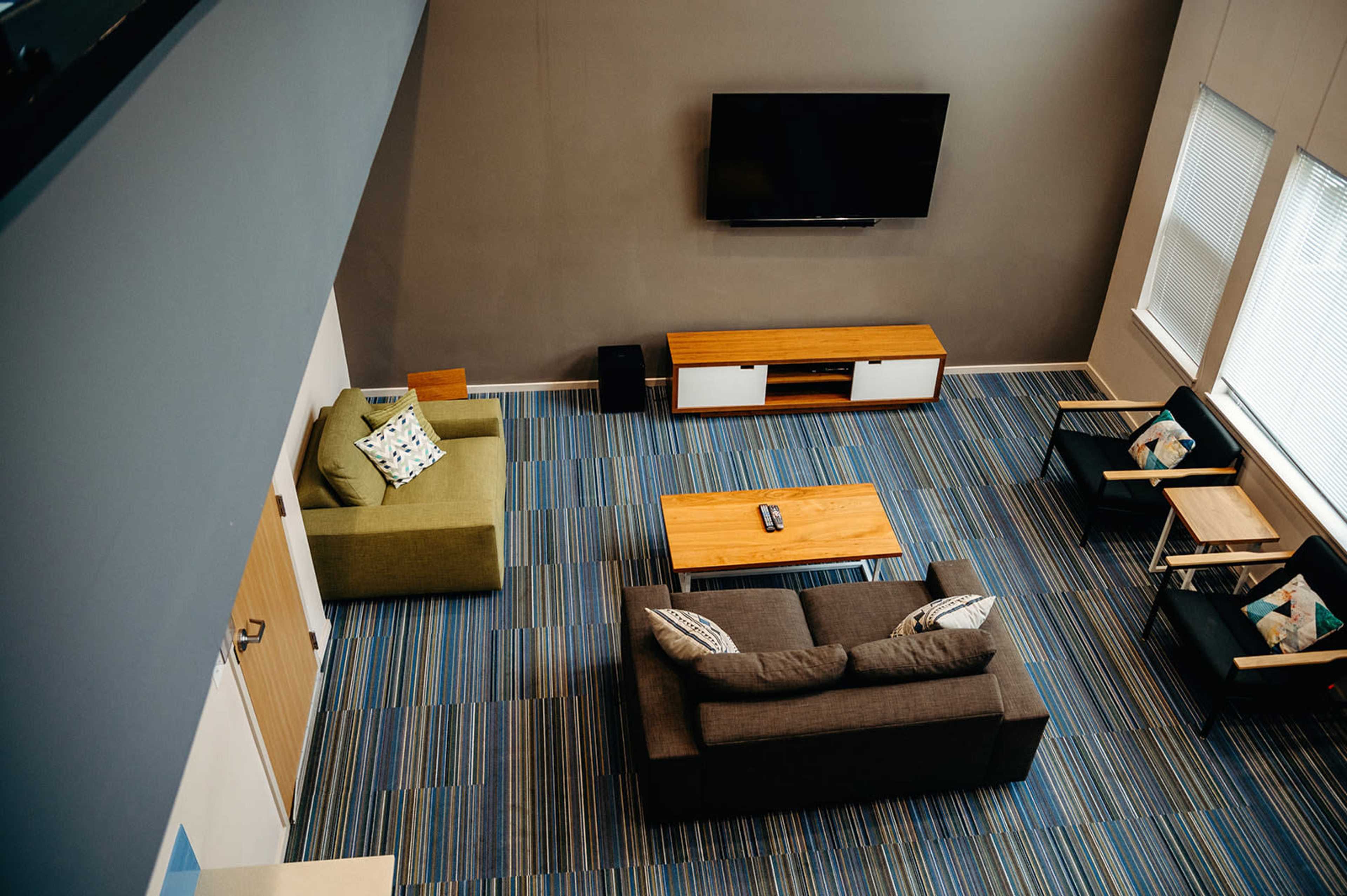 A modern living room features a dark gray sofa, a light wood coffee table, and two chairs on a striped blue carpet, with a television mounted on the wall.