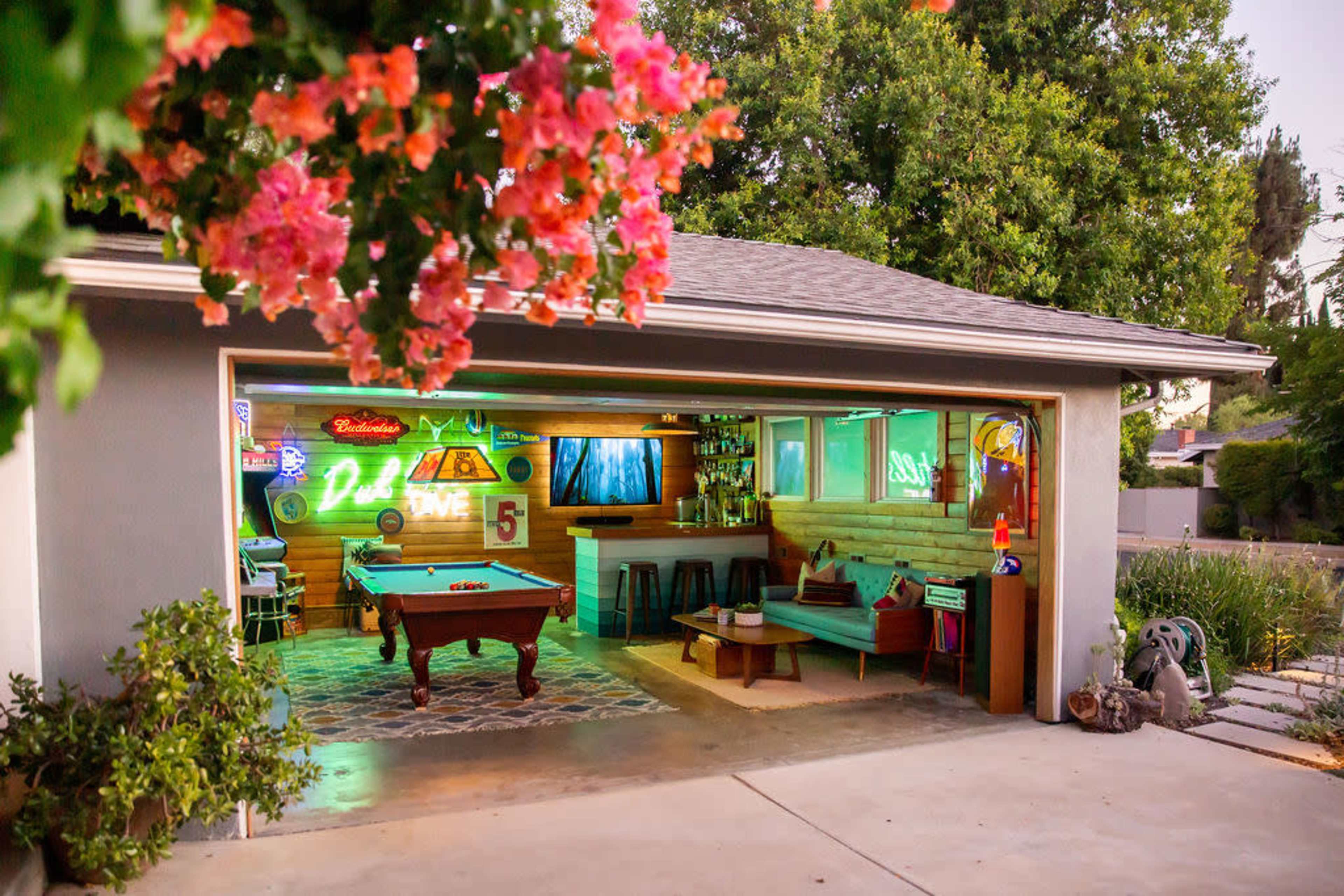 A garage converted into a recreational space with a pool table, neon lights, and a cozy seating area, surrounded by greenery.