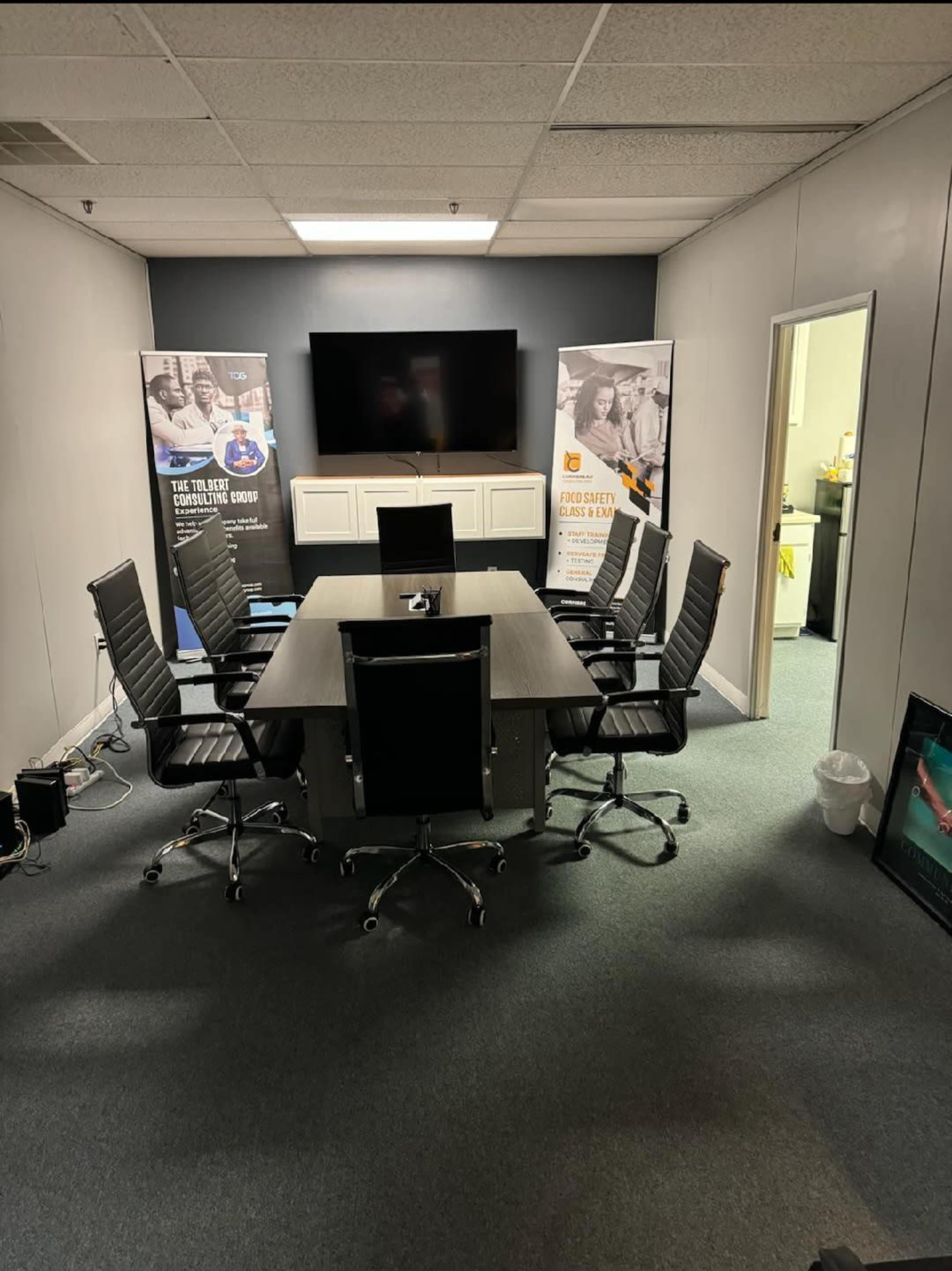 The image shows a modern conference room with a large table surrounded by six black office chairs, two promotional banners, and a wall-mounted screen.