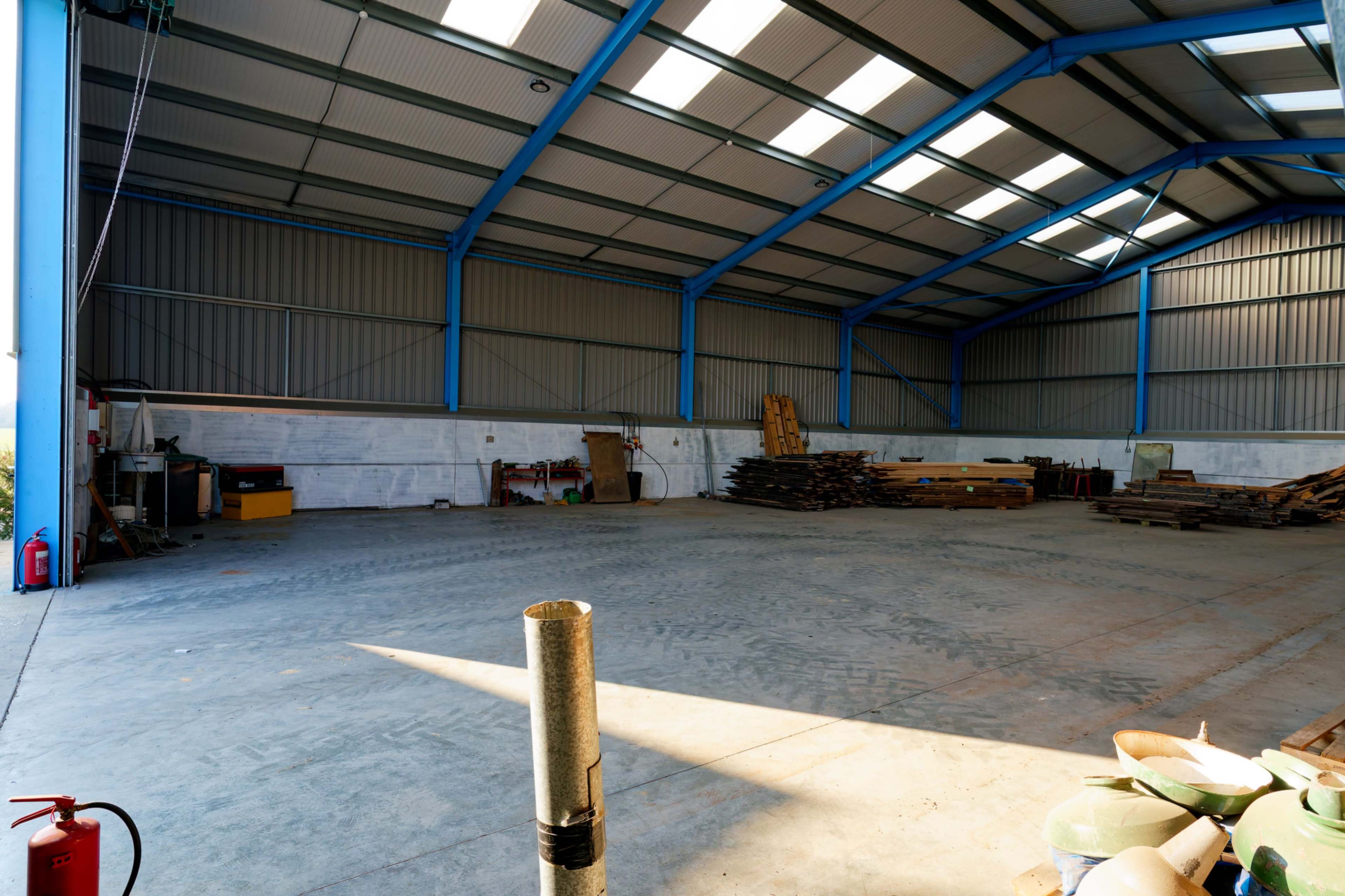 The image shows a spacious, empty warehouse with high ceilings, blue steel beams, and stacks of wooden materials along the back wall.