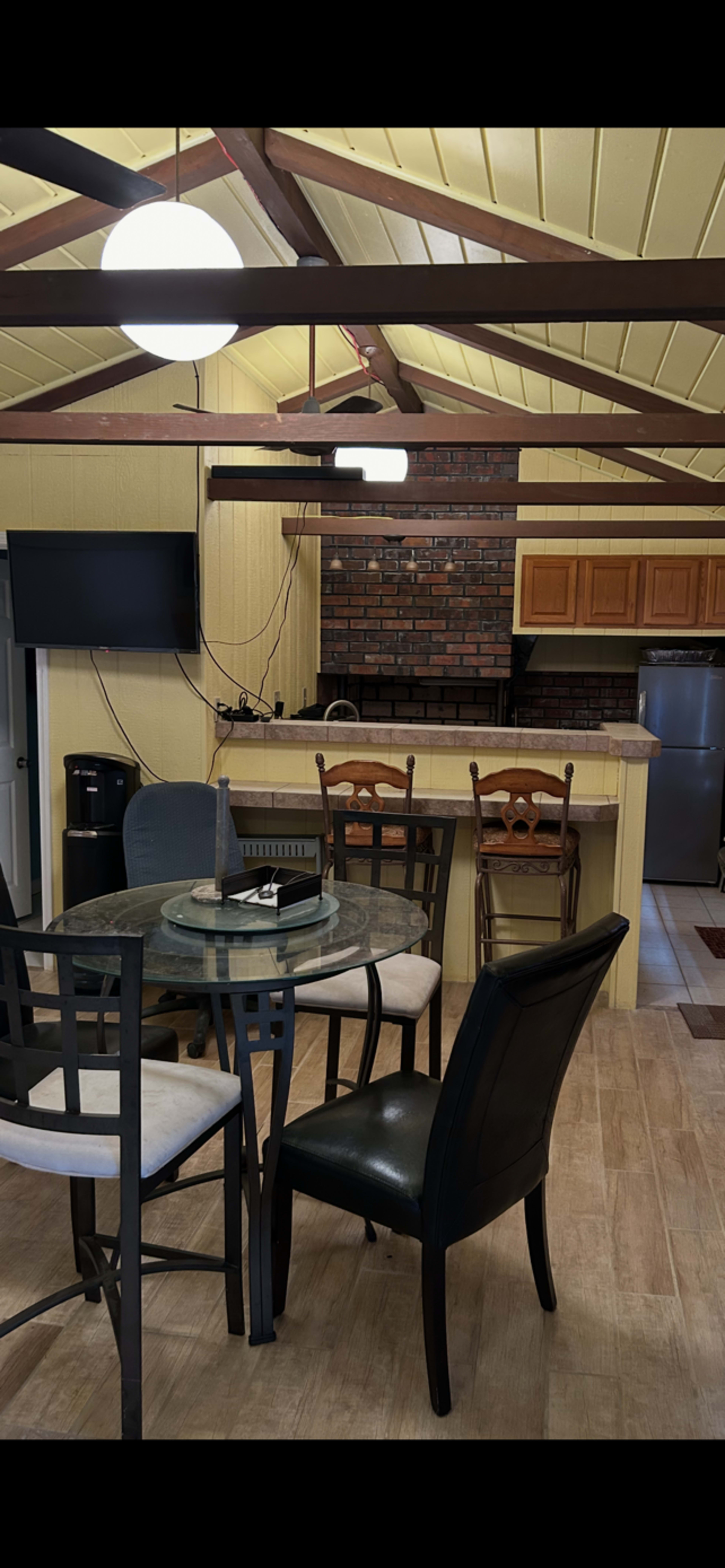 The image shows an interior view of a cabin-style kitchen and dining area with a circular glass table and several chairs, a bar counter with wooden cabinetry, and a brick backsplash.