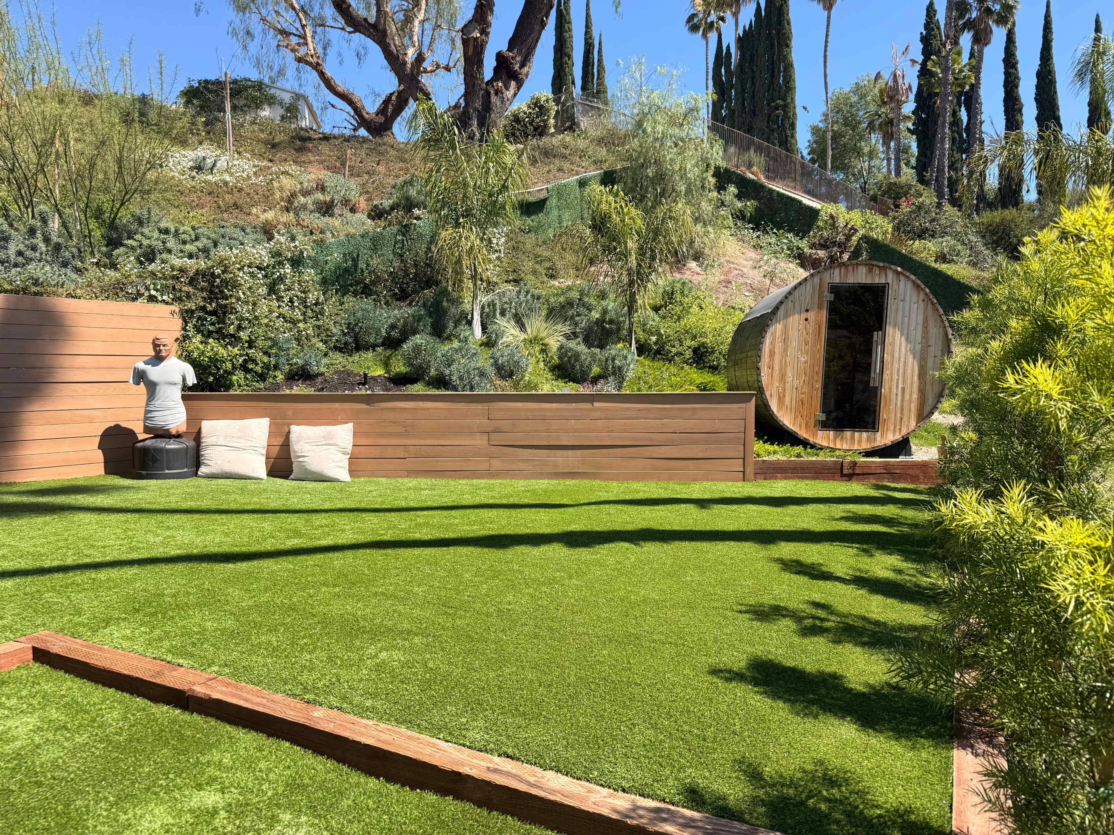 The image shows a landscaped outdoor area featuring artificial green grass, wooden seating, and a round wooden structure in the background.
