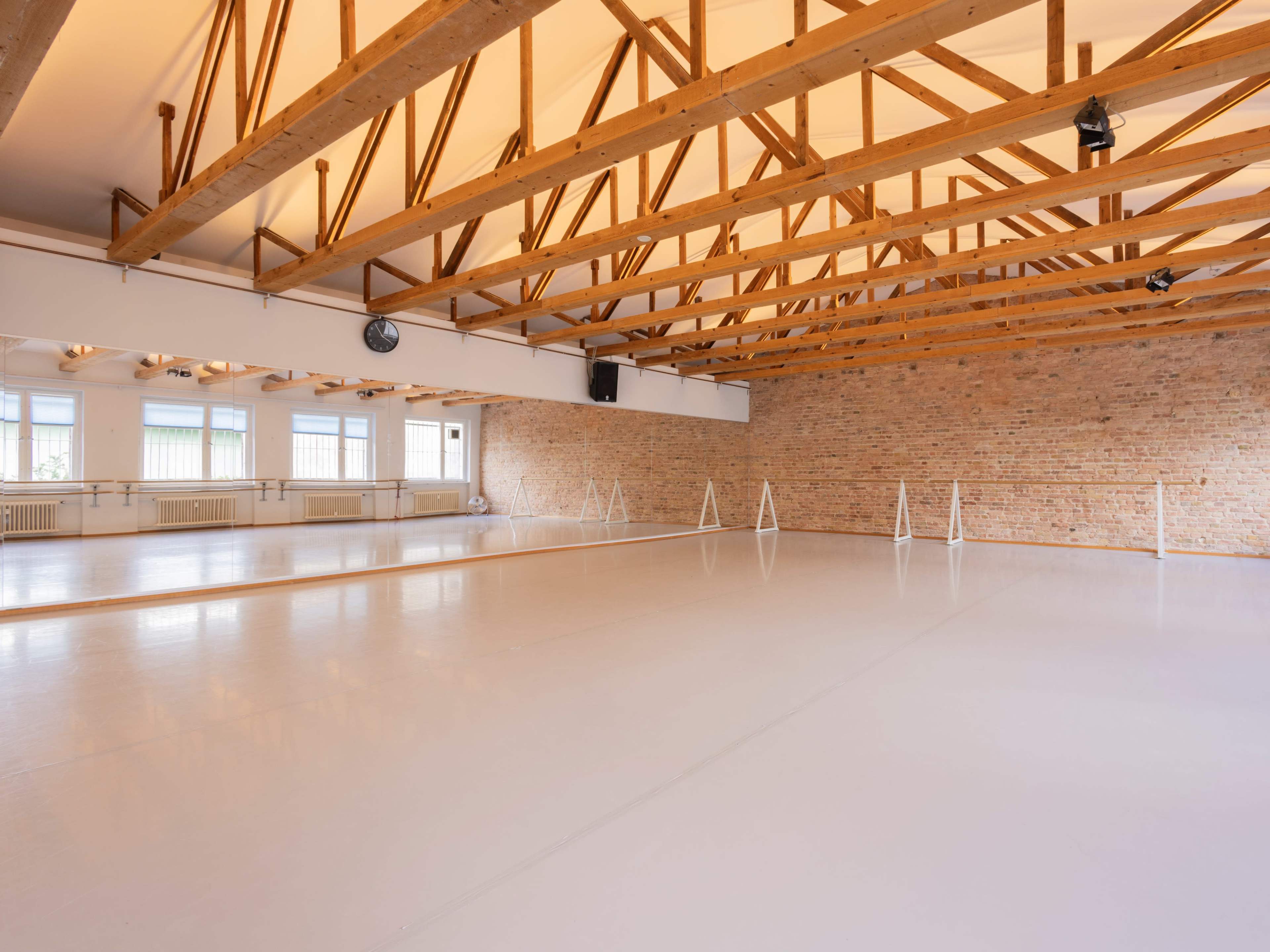 A spacious dance studio with wooden beams overhead, large windows allowing natural light, and a smooth floor with ballet barres along the walls.
