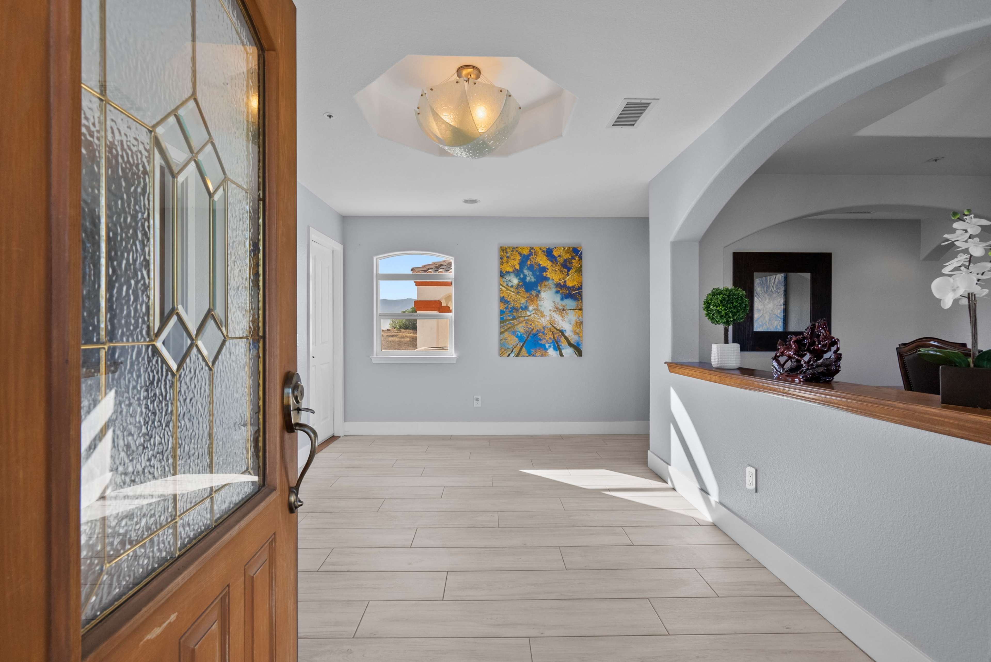 A well-lit entryway featuring a decorative door, tiled flooring, and a view of a living area with artwork on the walls.