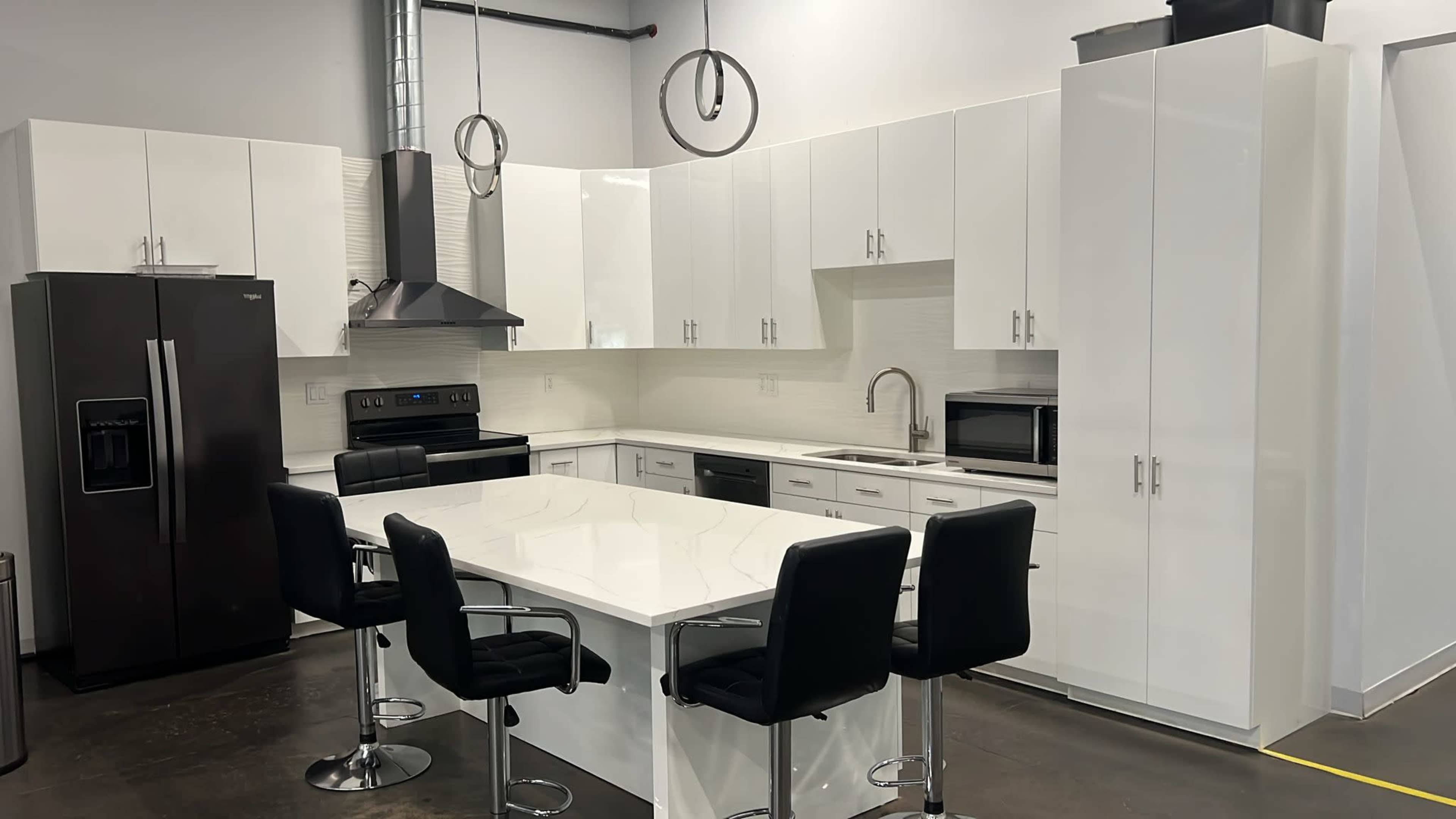 The image shows a modern, minimalist kitchen featuring white cabinetry, a black refrigerator, and a central island with barstool seating.