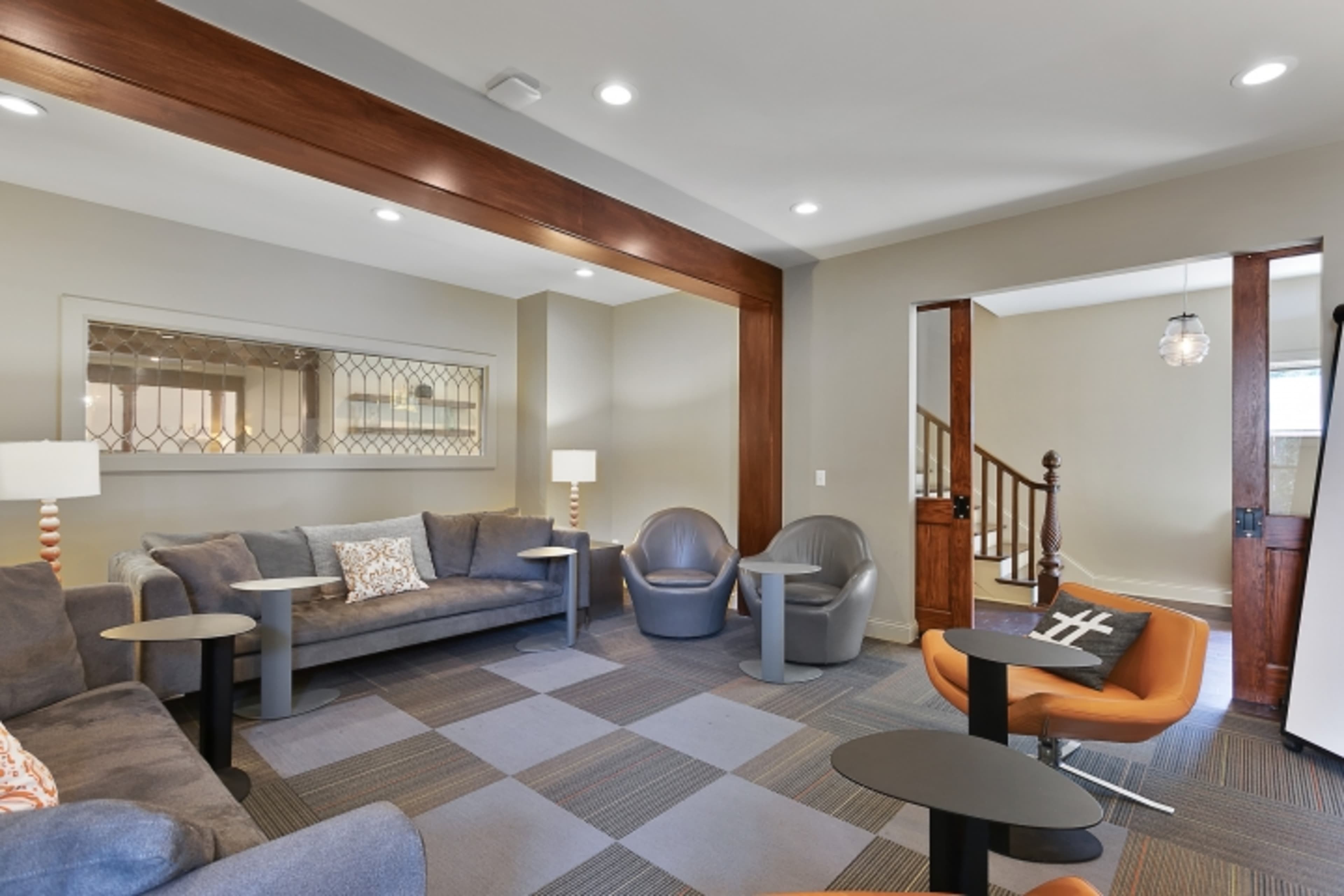 A modern lounge area with gray couches, armchairs, and small tables, featuring a decorative mirror and stairs leading to another level.