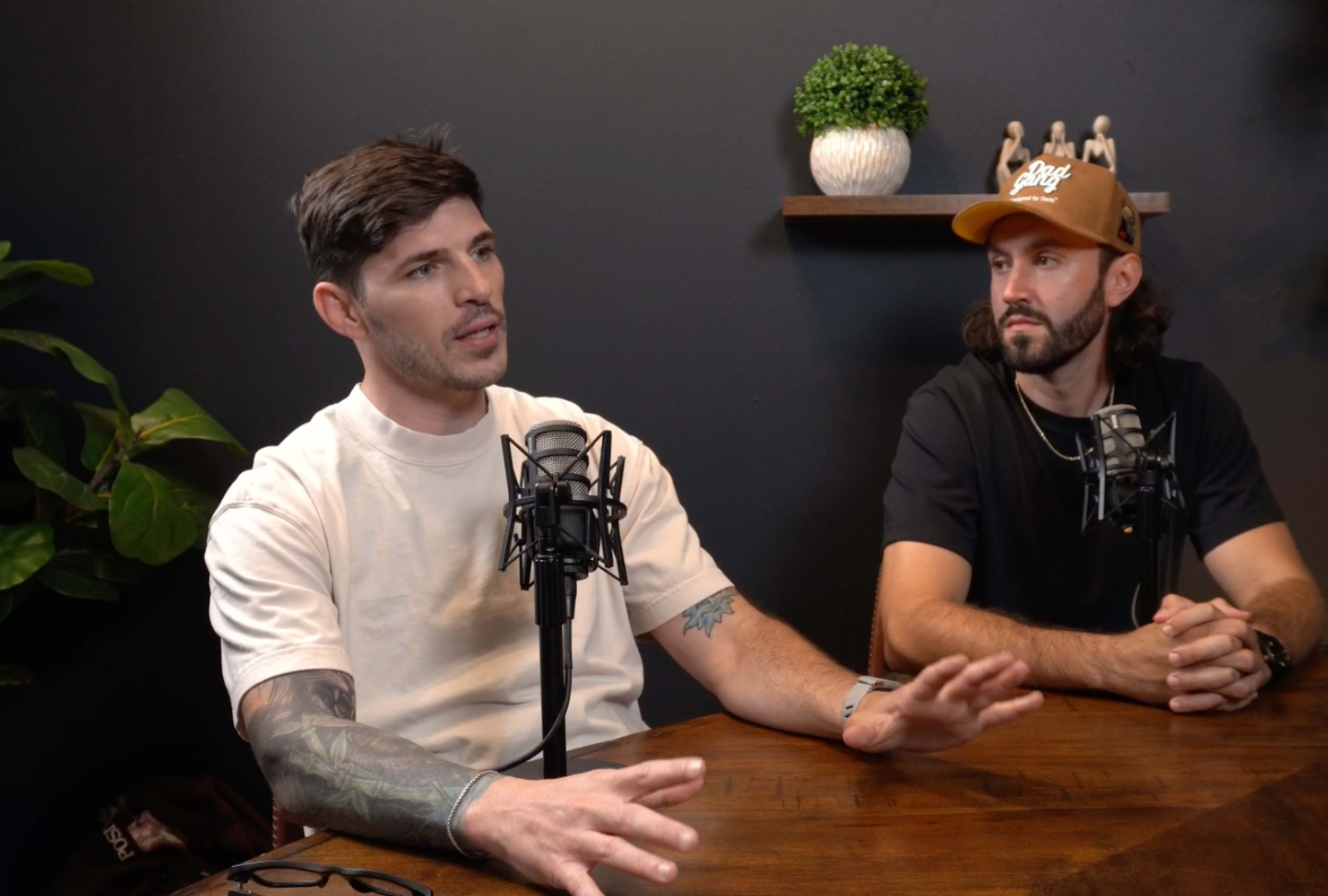 Two men are seated at a wooden table in a studio setting, discussing while microphone stands are positioned in front of them.