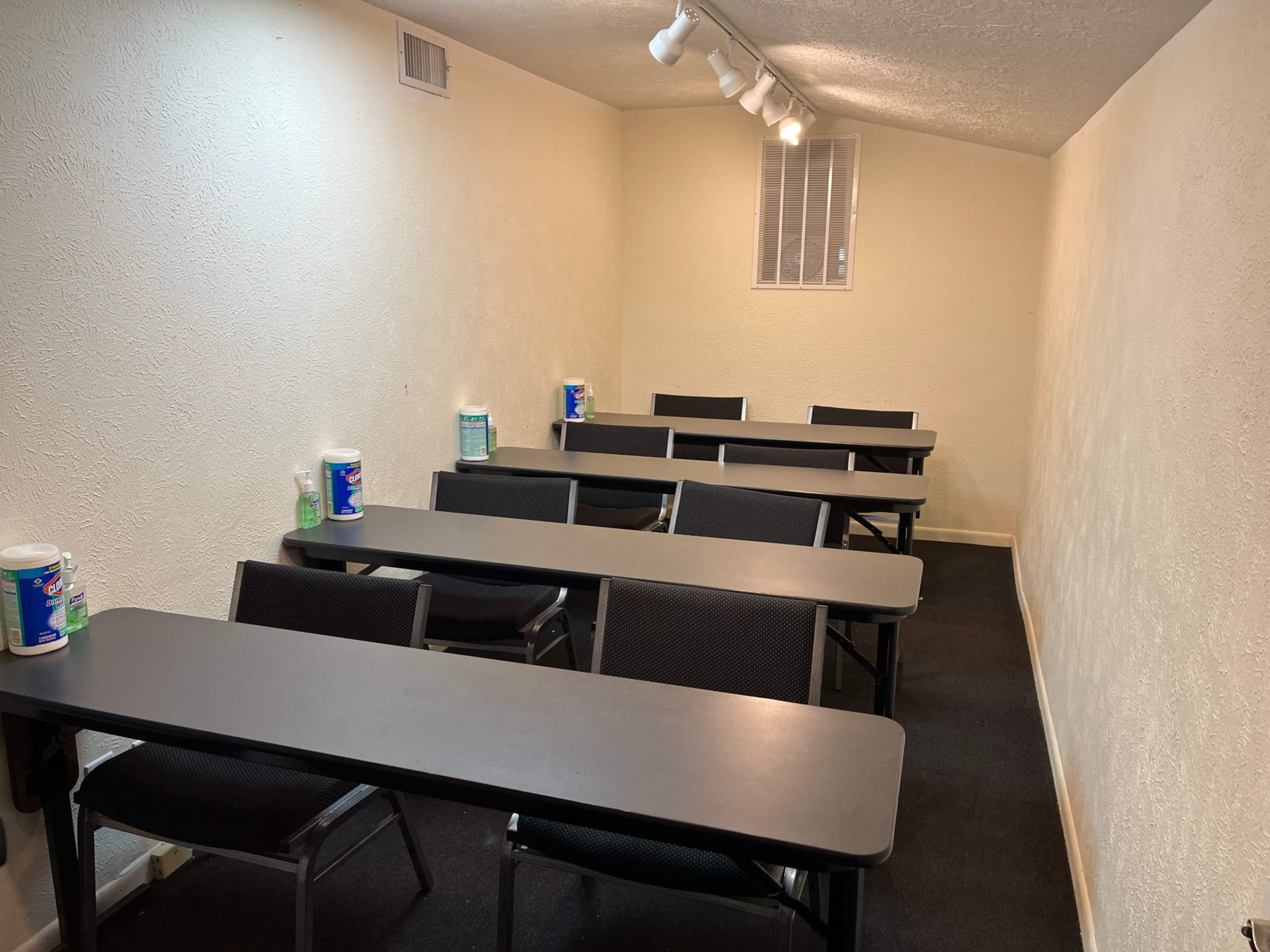 A narrow classroom features several rows of black tables and chairs positioned on a dark carpeted floor, with disinfectant supplies placed on the tables.