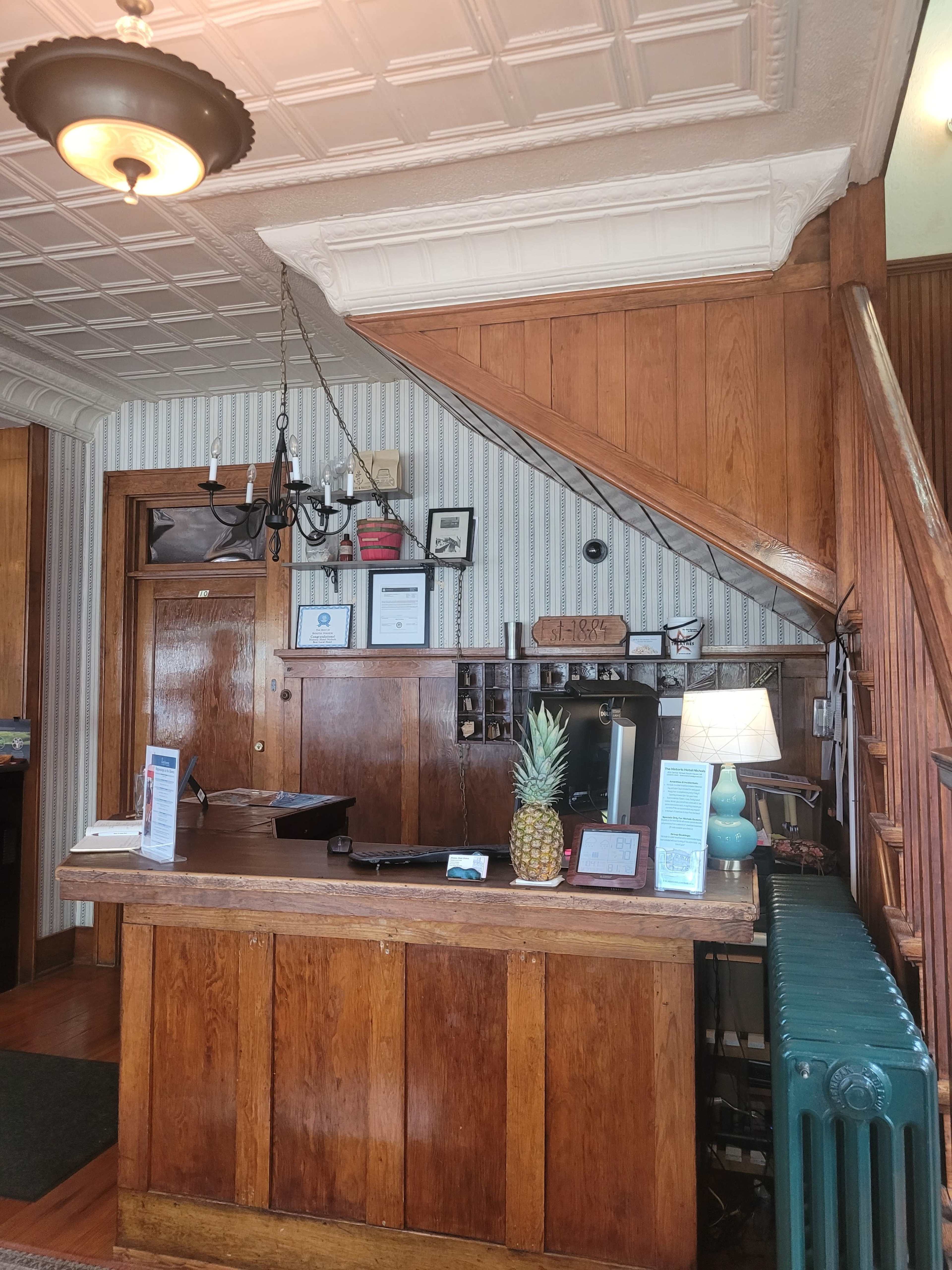 A wooden reception desk with a pineapple centerpiece, located in an area featuring a staircase and wall-mounted decor.