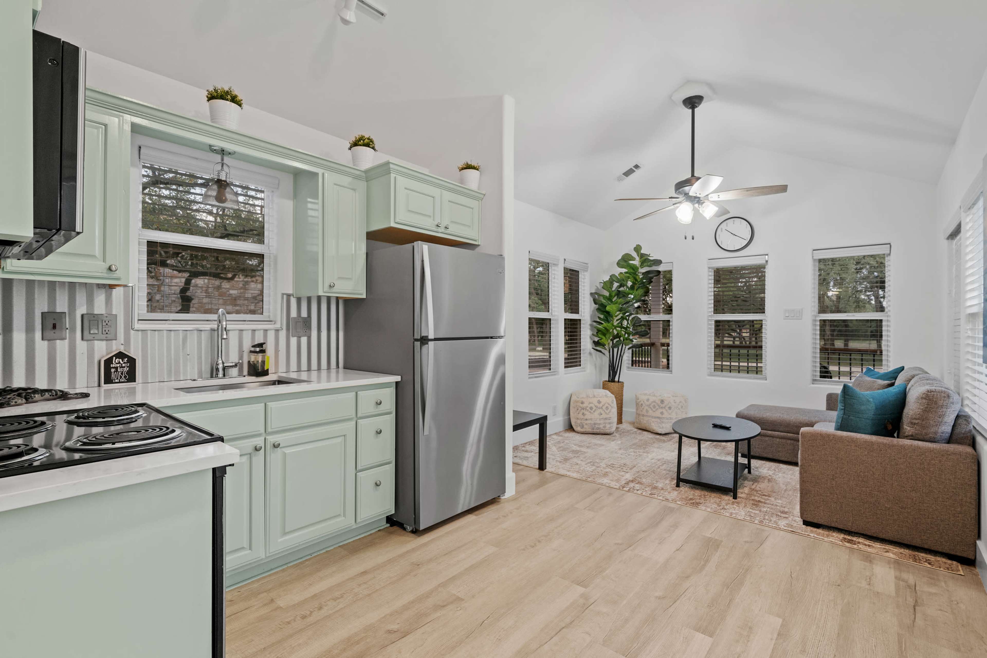 A modern kitchen with light green cabinetry and a stainless steel refrigerator opens up to a bright living area featuring a gray sofa and large windows.
