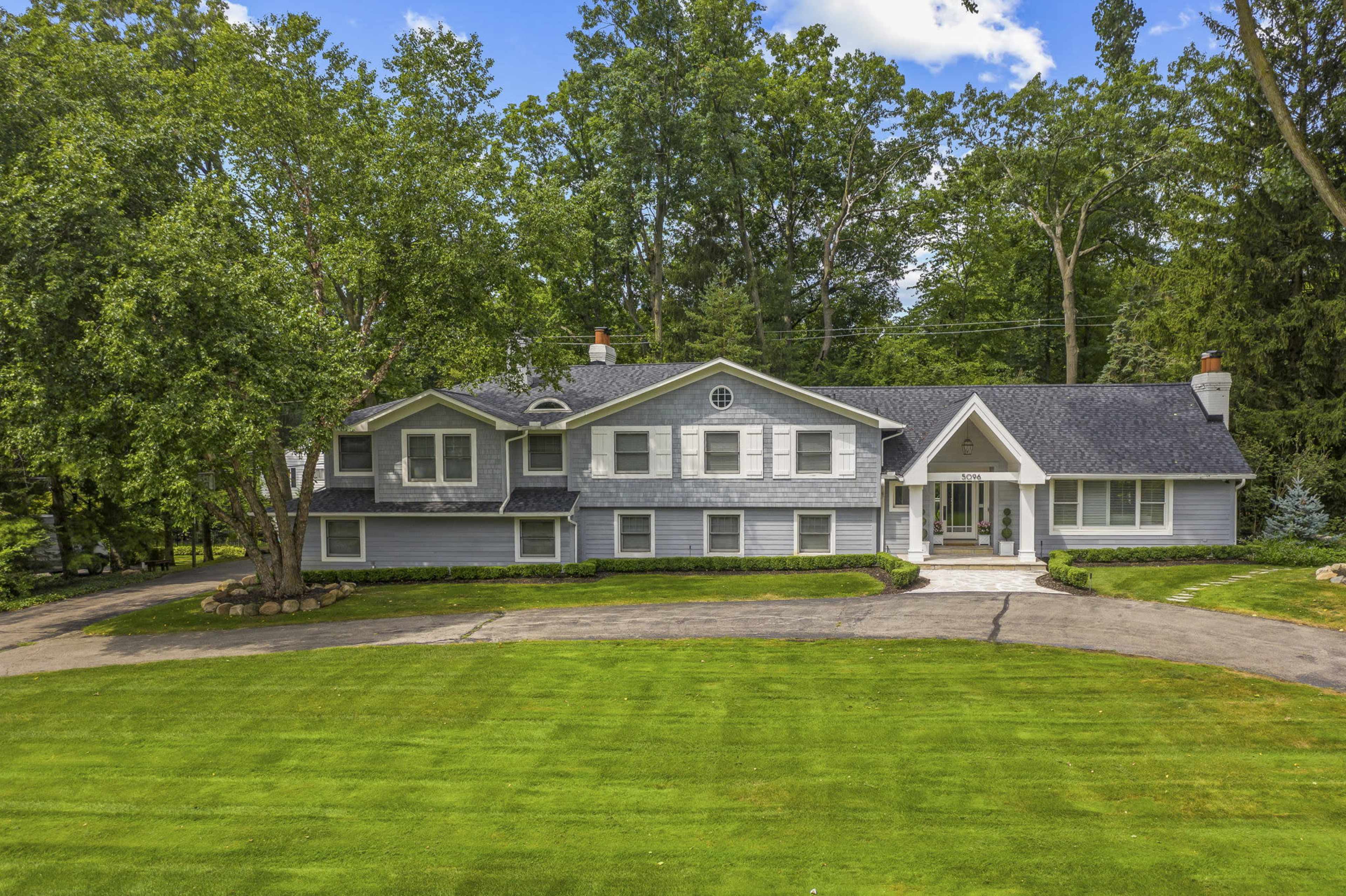 The image shows a large, two-story gray house with multiple windows situated on a manicured lawn surrounded by trees.
