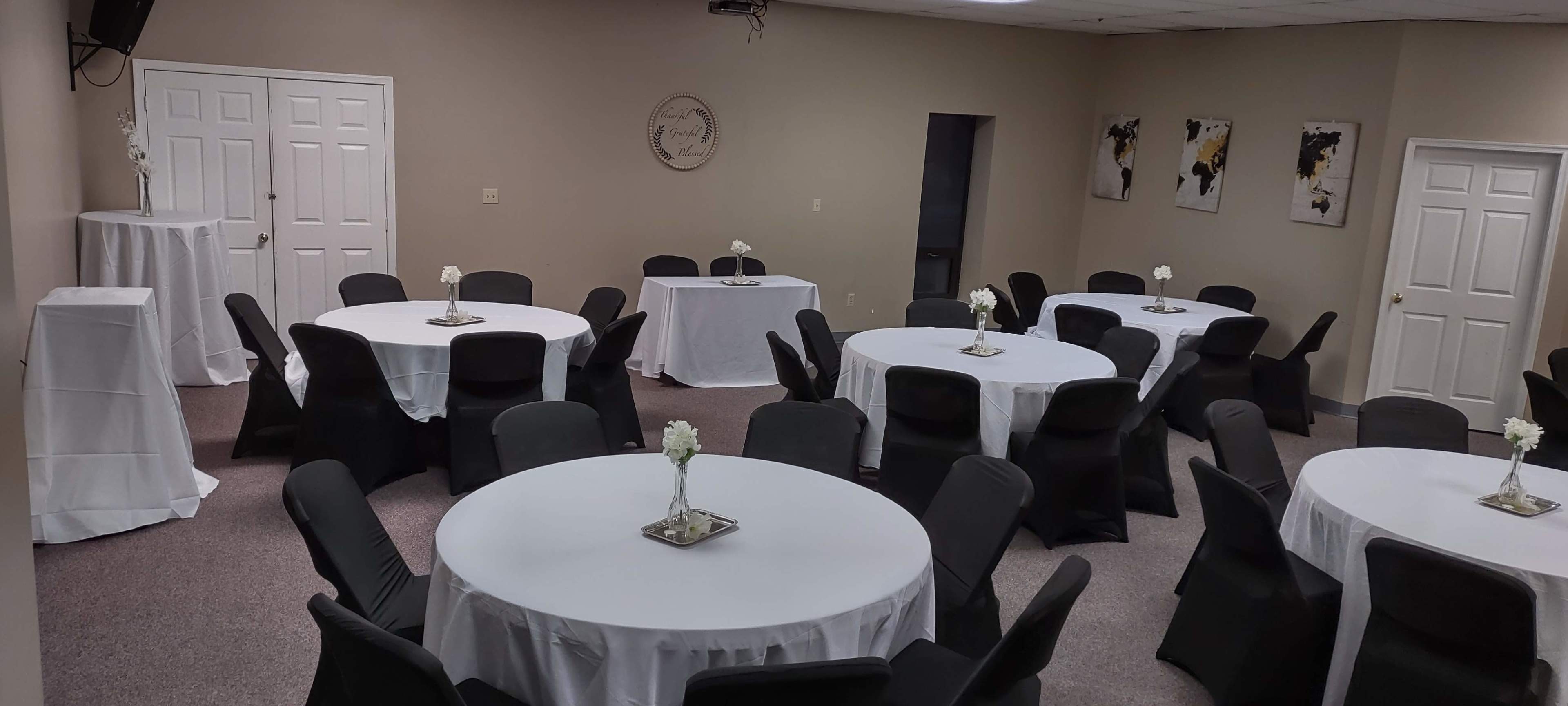 A meeting room set up with several round tables covered in white tablecloths and black chairs, arranged around a central area.