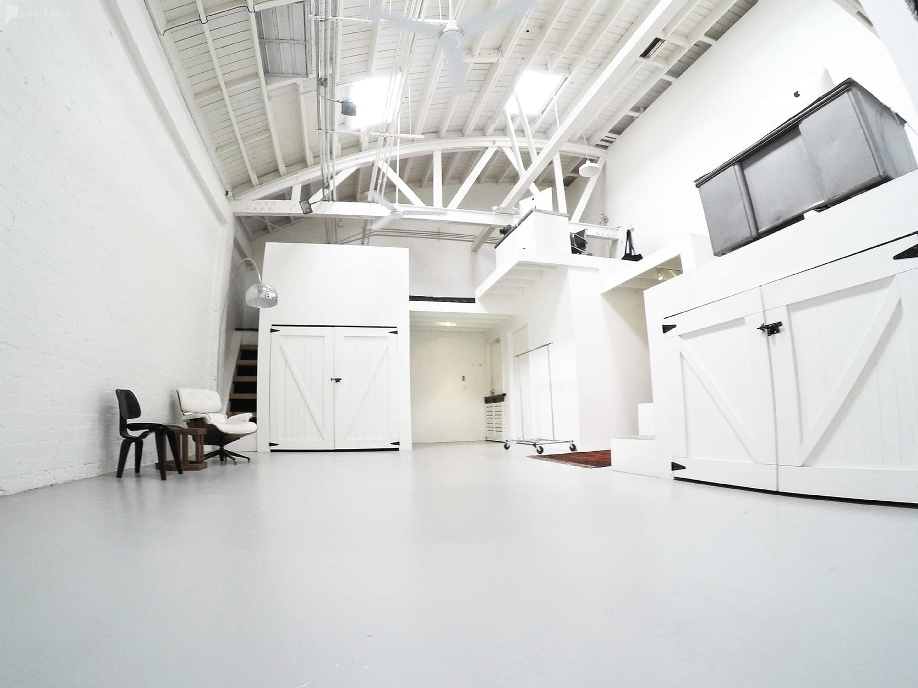 A spacious, modern interior with white walls, light flooring, and minimalist furnishings, including a black chair and a staircase leading to an upper level.