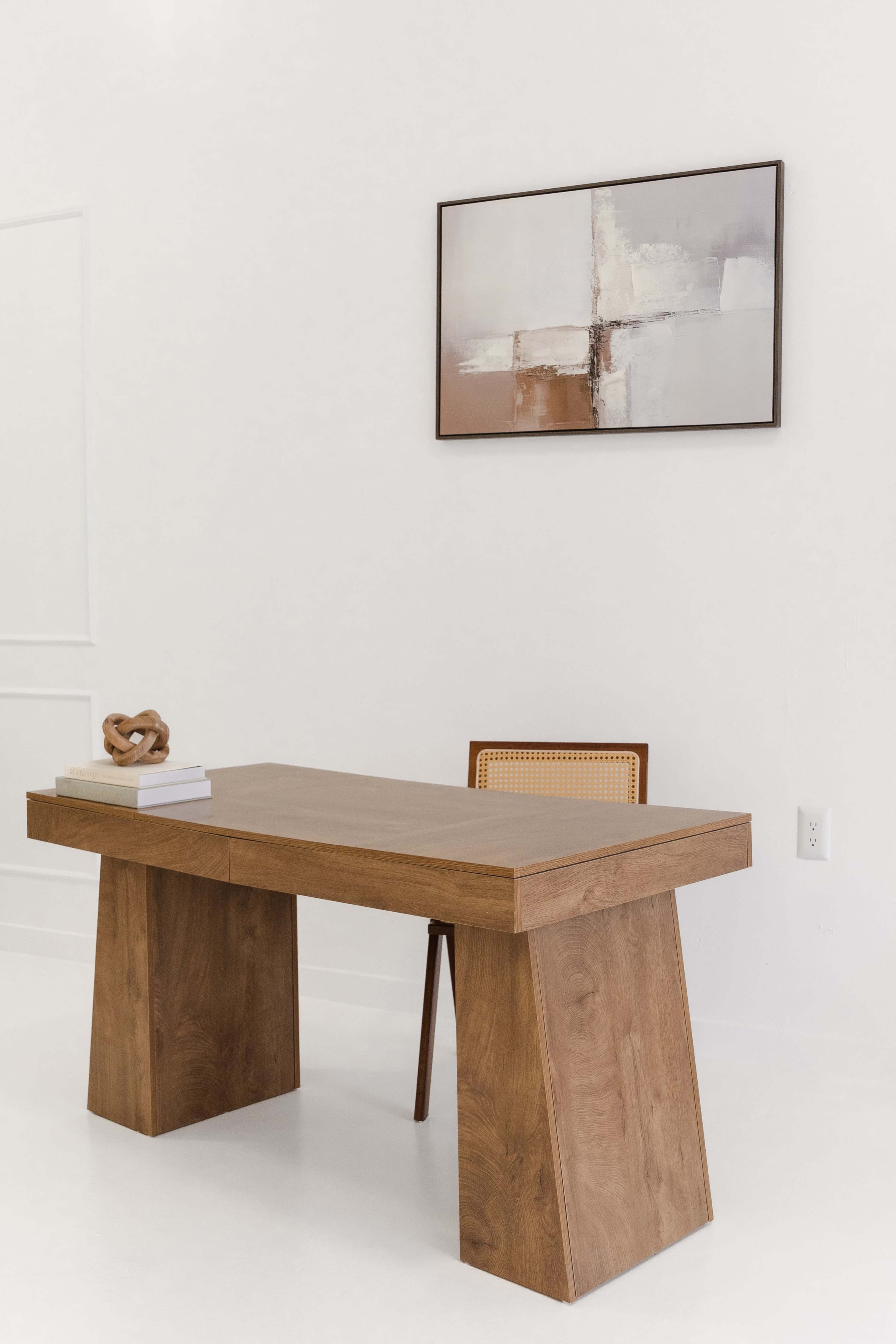 The image shows a minimalist workspace featuring a wooden desk with a geometric base, a framed abstract painting on the wall, and a simple chair.