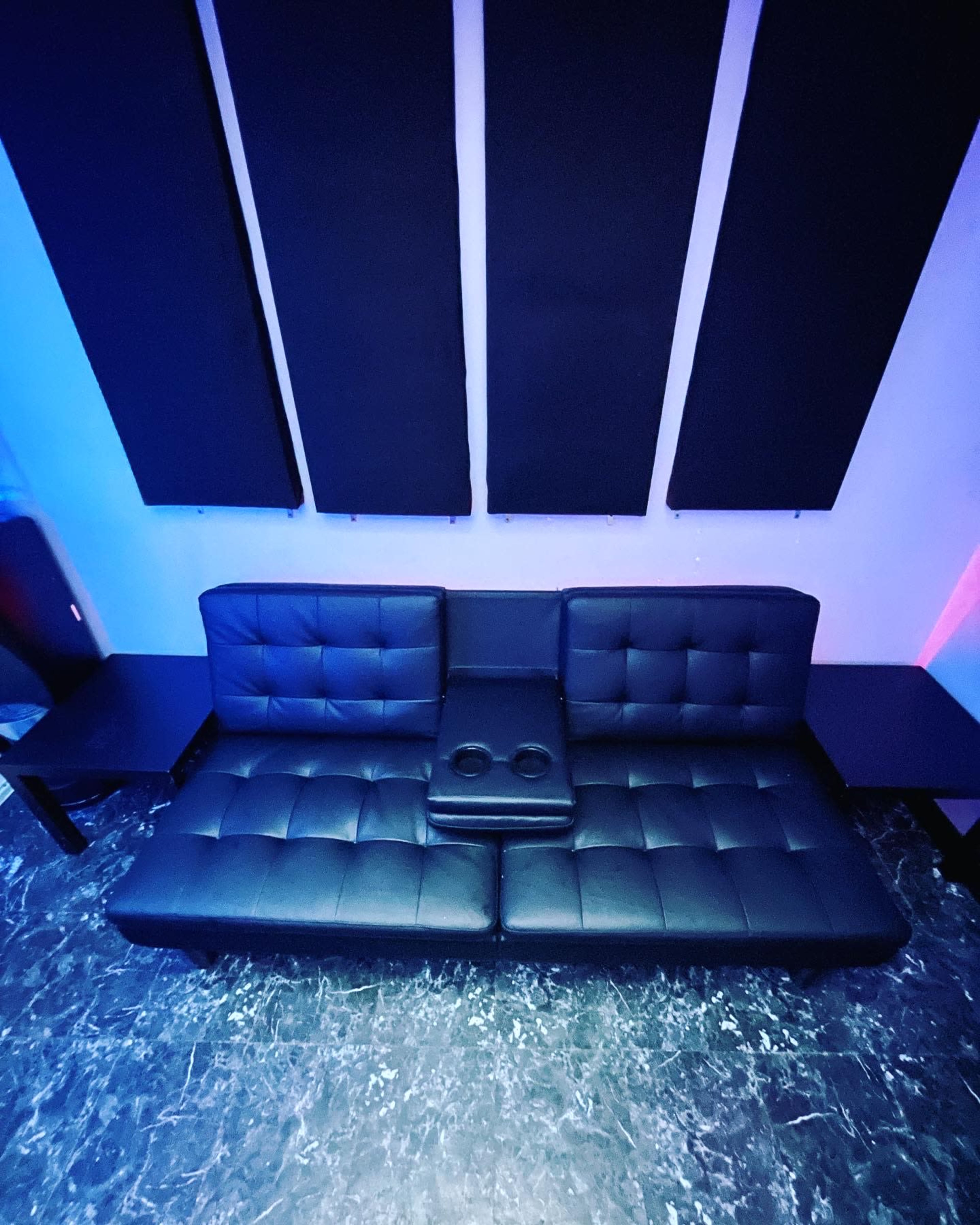 A black leather couch with a central cup holder, positioned between two small tables against a backdrop of soundproof panels.