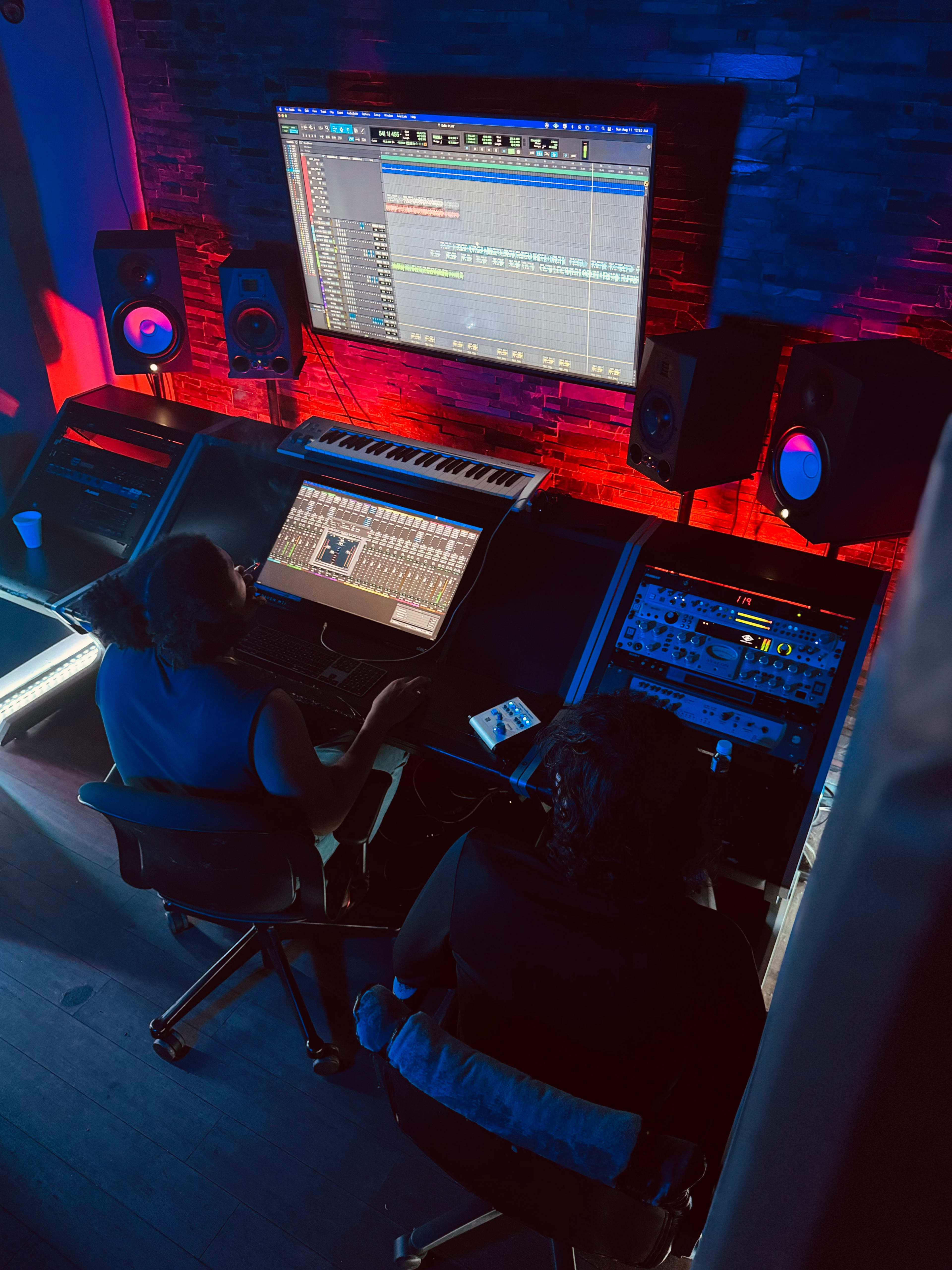 Two individuals are working on music production in a recording studio, focused on a computer screen displaying audio tracks and equipped with various sound equipment and a keyboard.