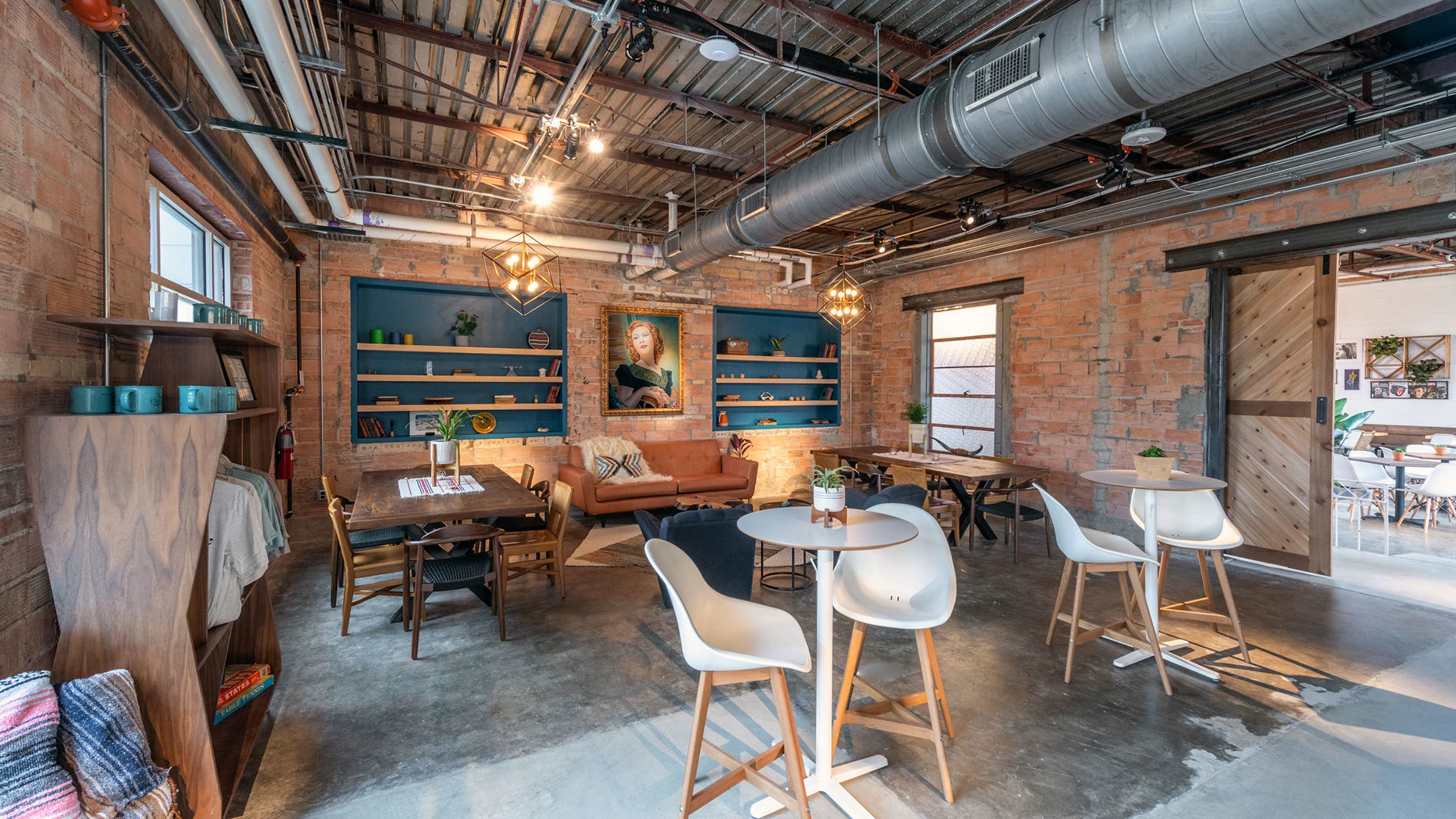 A modern lounge area features exposed brick walls, wooden shelving, and a mix of seating arrangements, including tables and a cozy couch.