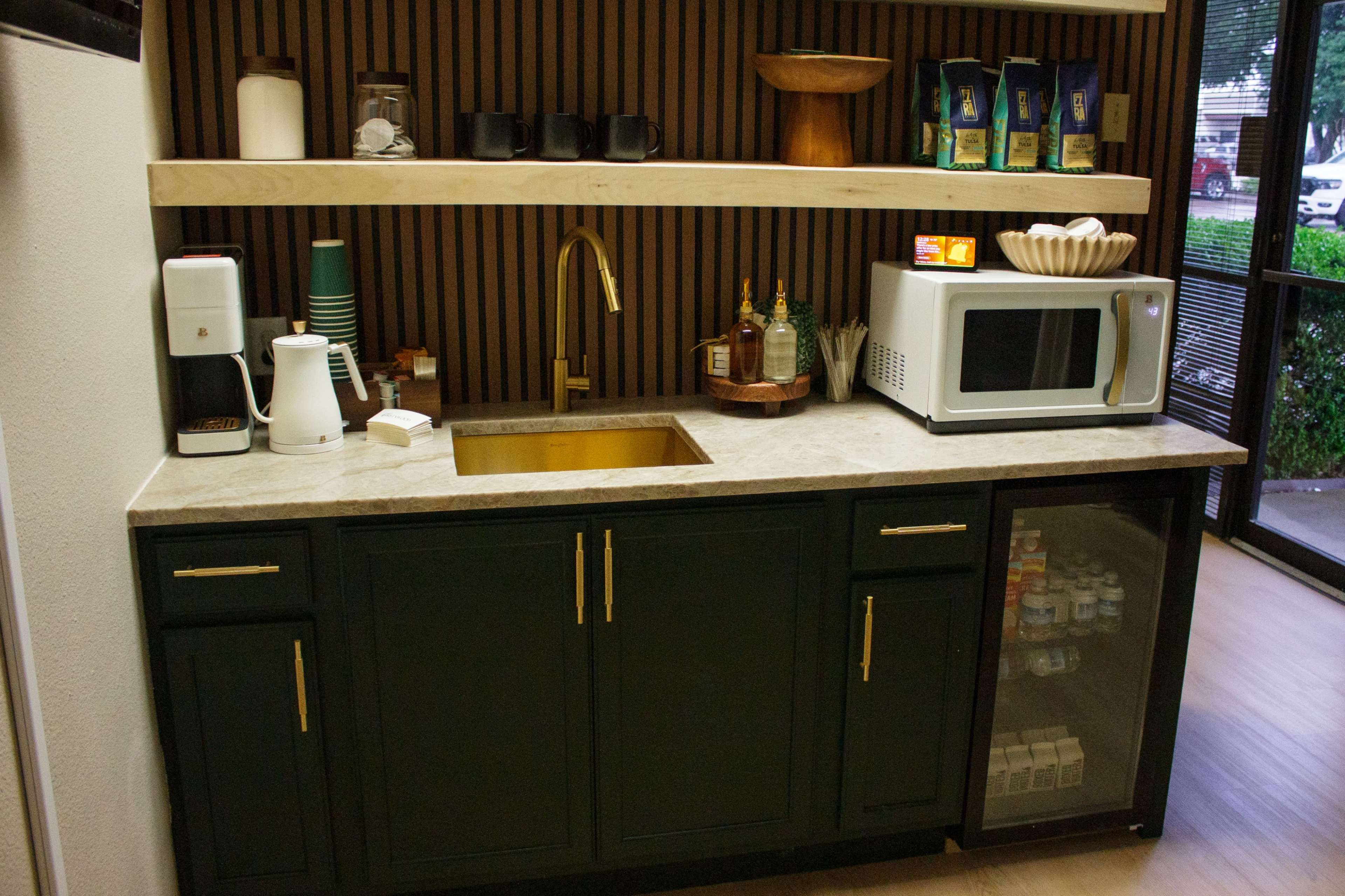 The image shows a kitchenette area with a dark green cabinet, a microwave, a gold faucet above a sink, and a countertop displaying various items including coffee cups and bottles.