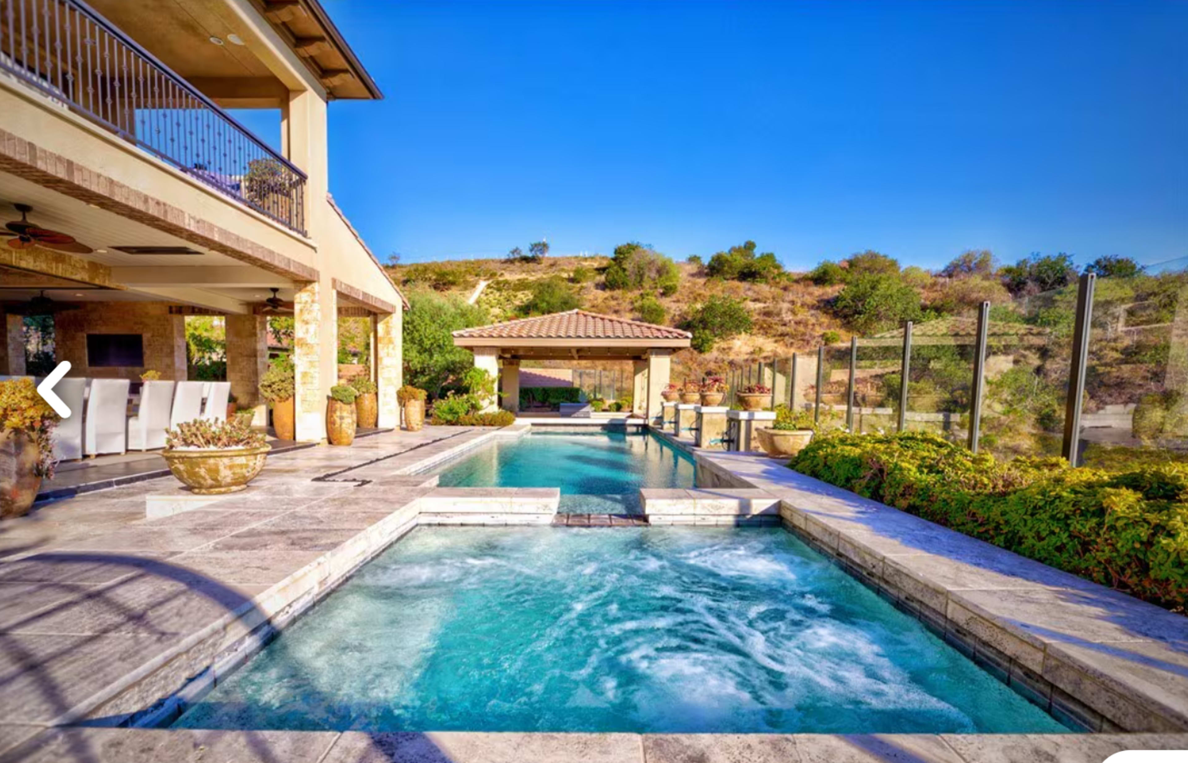 The image shows a serene backyard featuring a swimming pool and spa, surrounded by landscaped greenery and a hillside in the background.