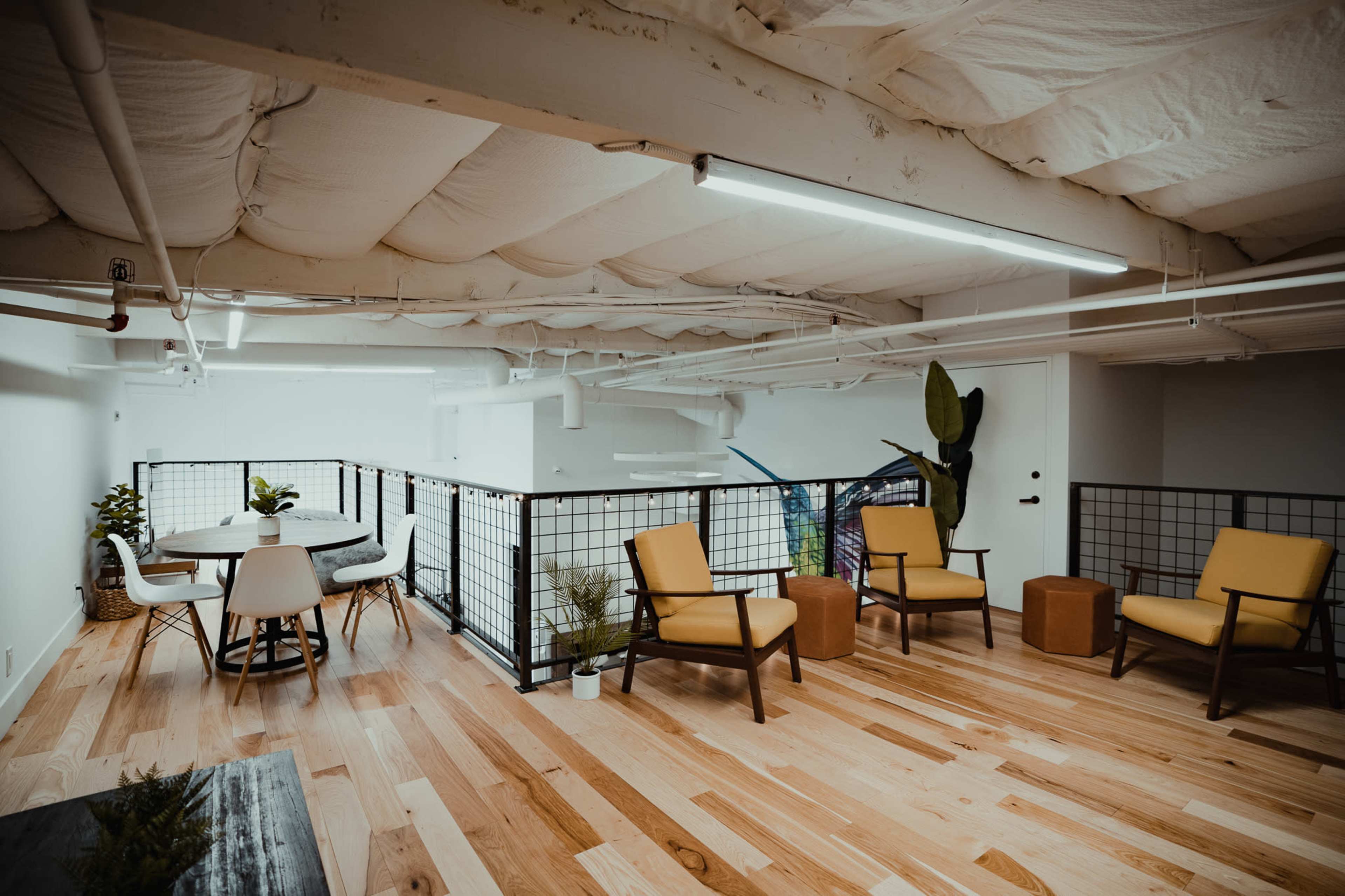 A modern office space with wooden flooring, featuring a seating area with yellow chairs, a round table, and decorative plants.