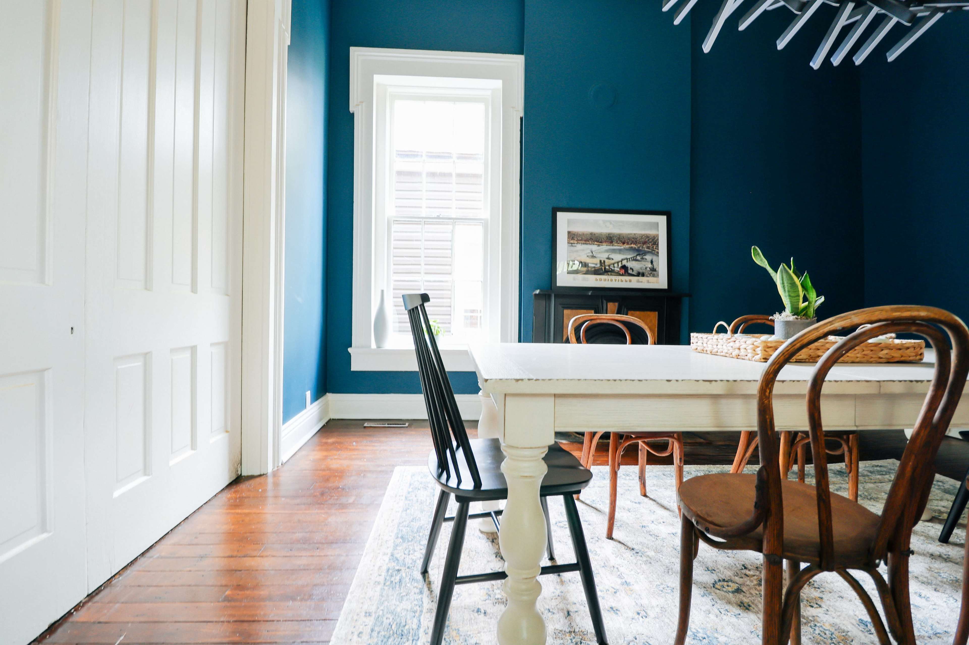 A dining area with a white table surrounded by wooden chairs, set against deep blue walls and a large window letting in natural light.