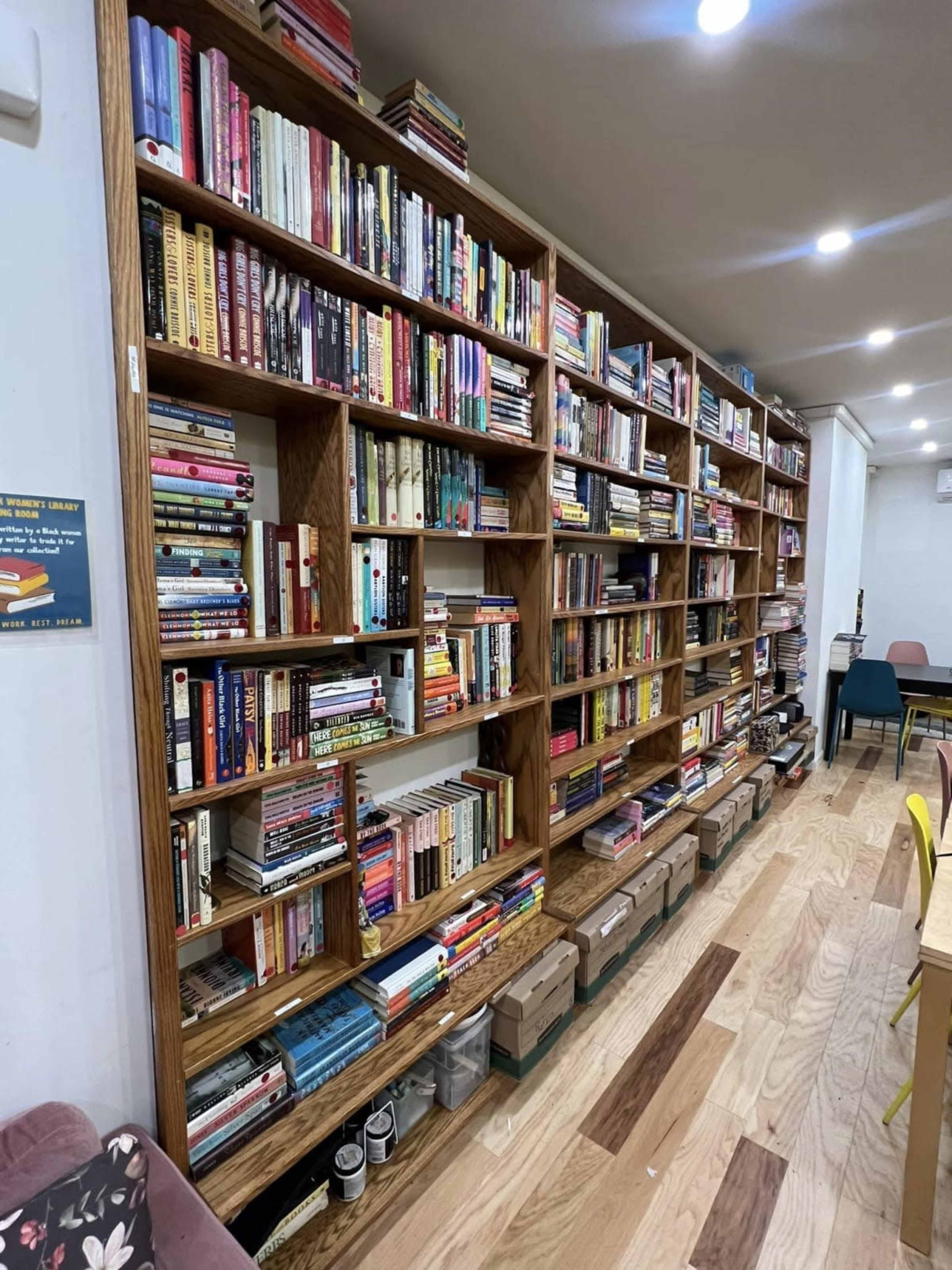 A wall-mounted bookshelf filled with various books and containers lines one side of a room with wooden flooring.