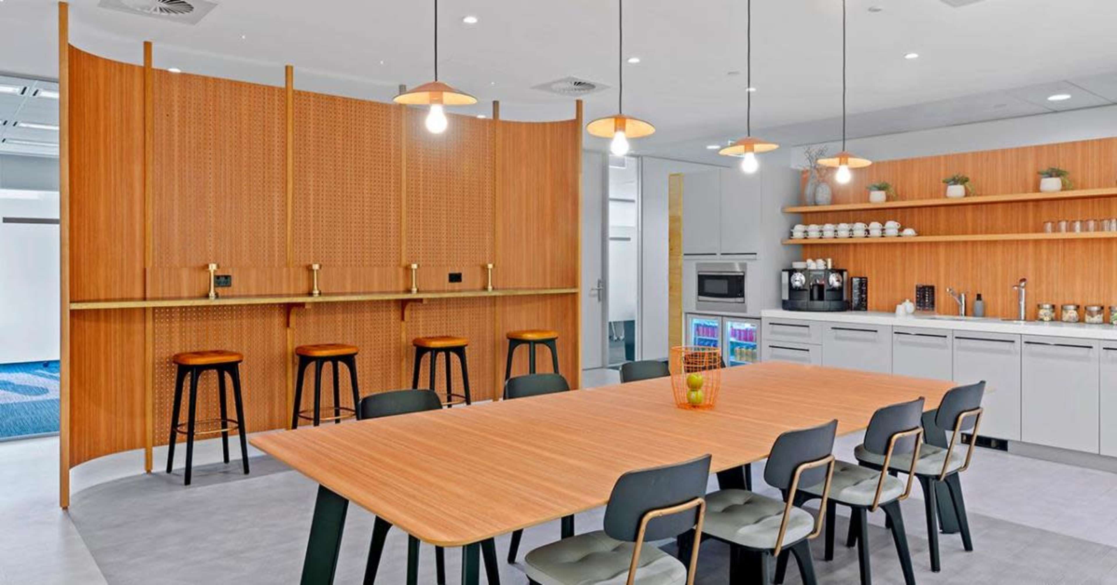 A modern kitchen and dining area with a large wooden table surrounded by chairs, a bar with stools, and appliances neatly arranged on shelves.