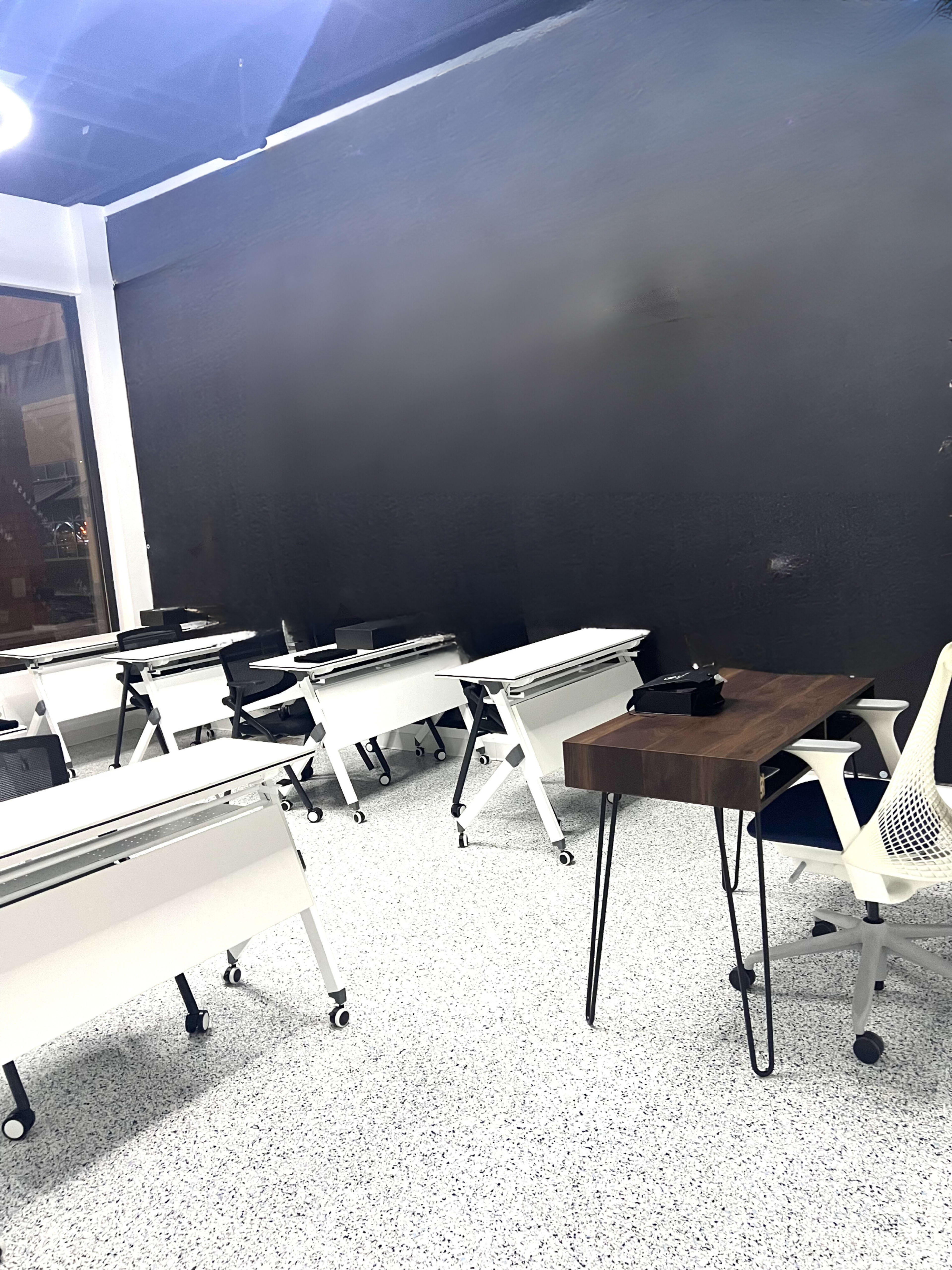 A classroom with several movable desks and a central wooden table, set against a dark wall.