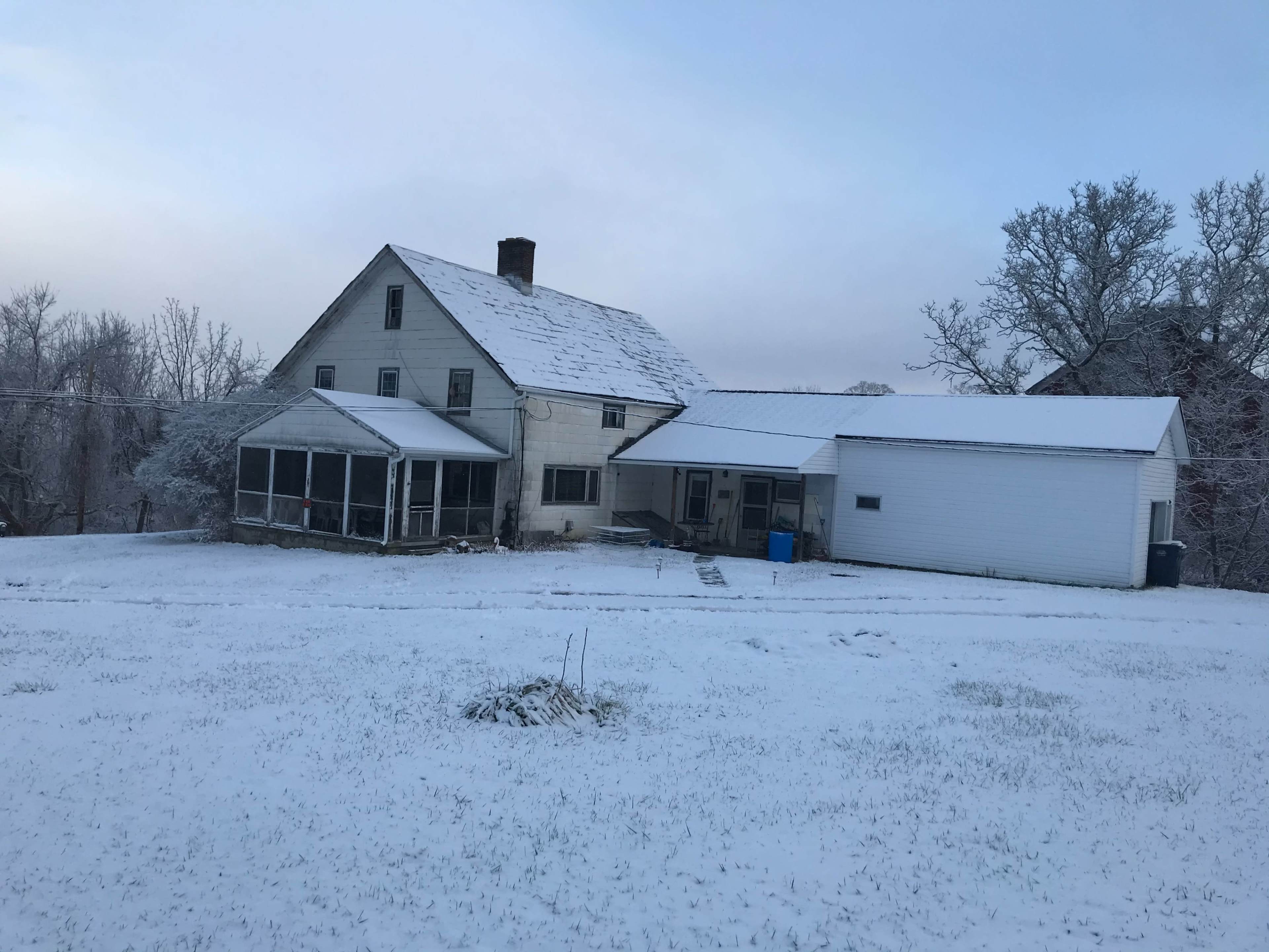A weathered house with a screened porch and an attached garage is surrounded by a blanket of snow.