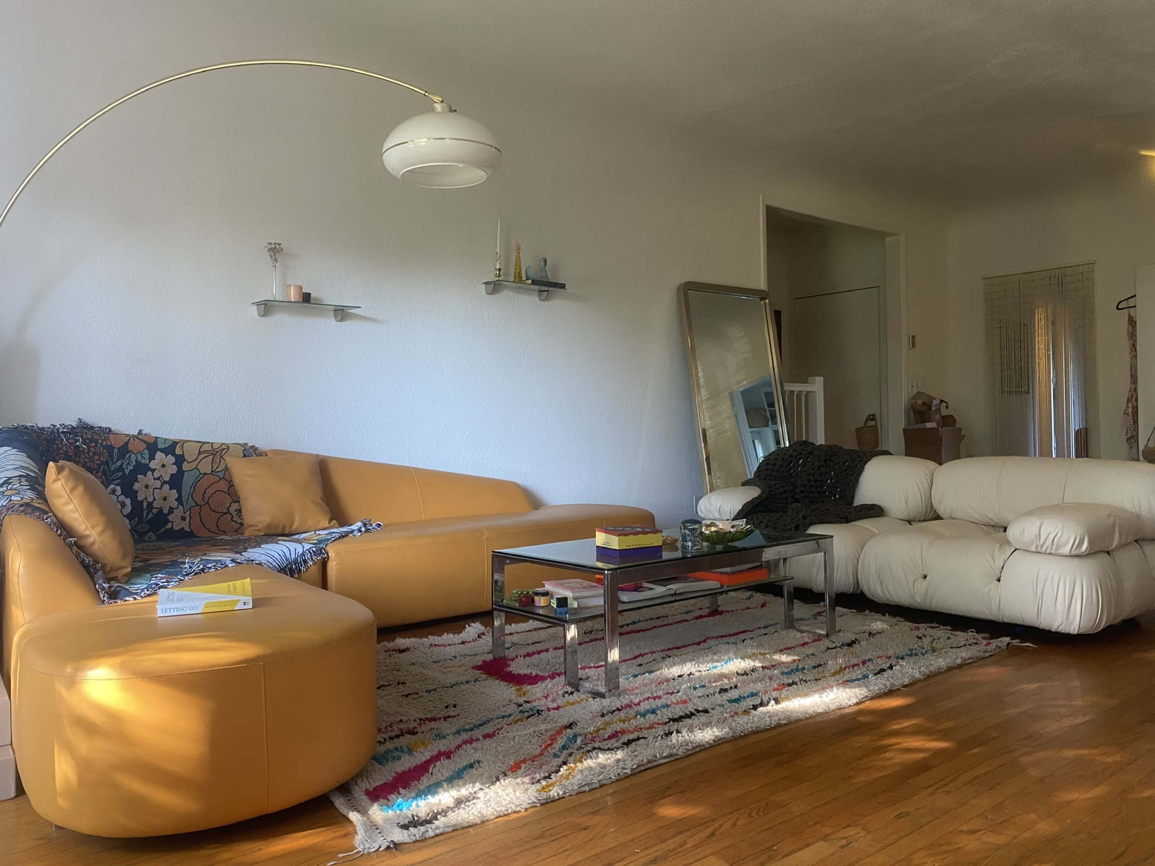A living room with a yellow sofa, a white lounge chair, a glass coffee table, and decorative items on a colorful rug.