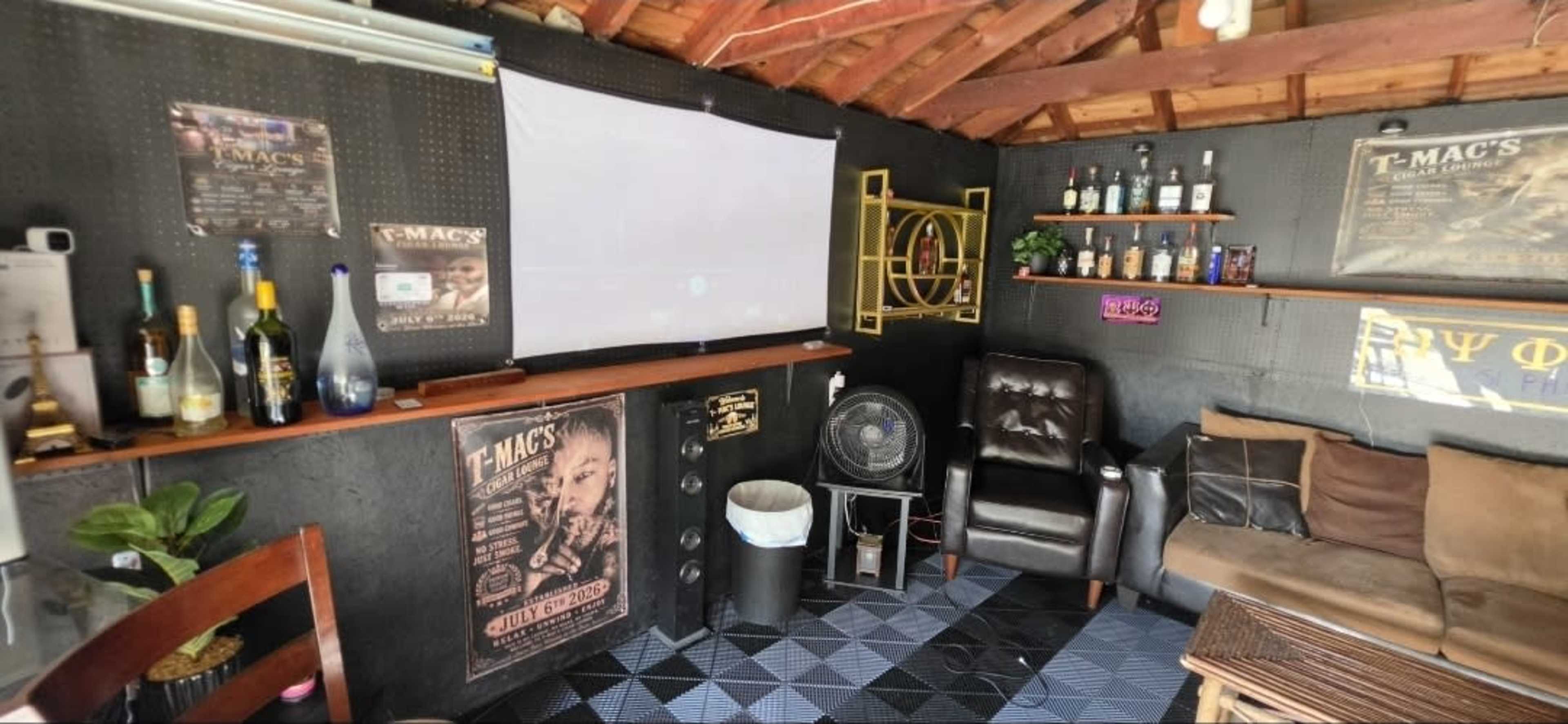 The image shows a cozy room with a large projection screen, a comfortable chair, a sofa, and shelves displaying various bottles and posters on the walls.
