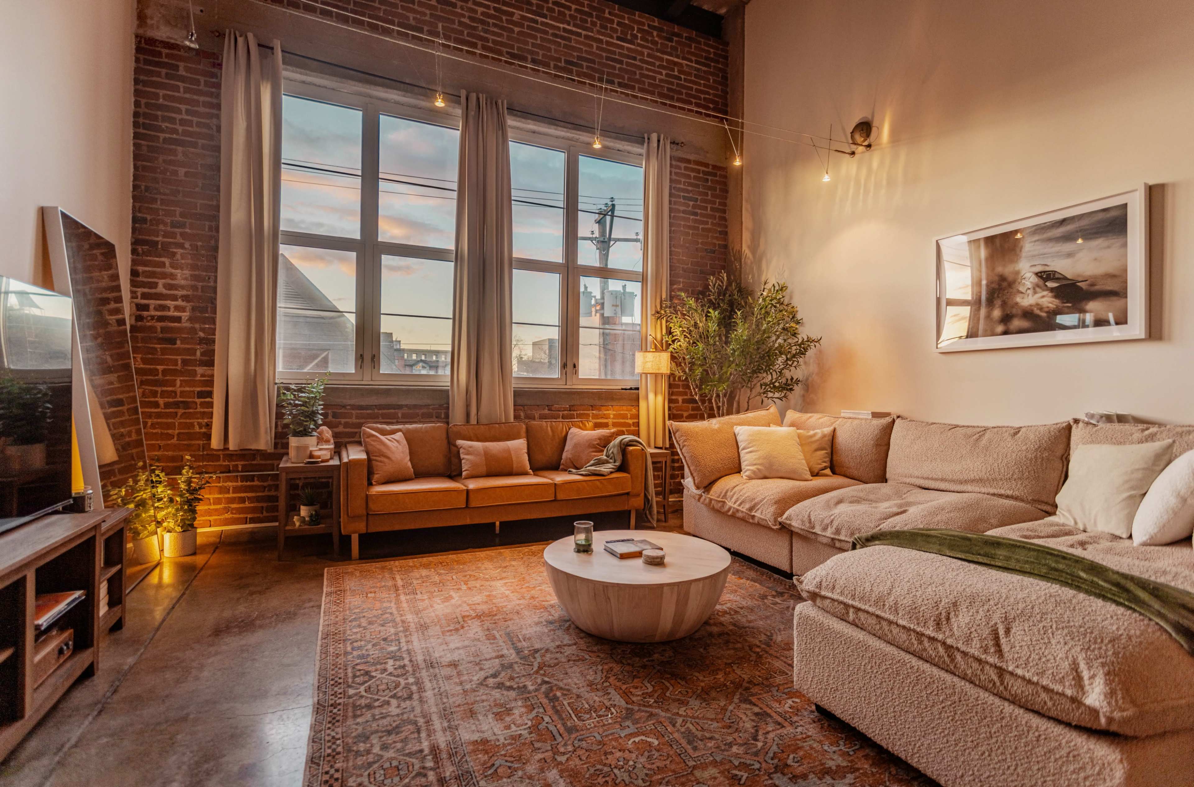 The image features a cozy living room with a large sectional sofa and a round coffee table, set against a backdrop of exposed brick walls and large windows.