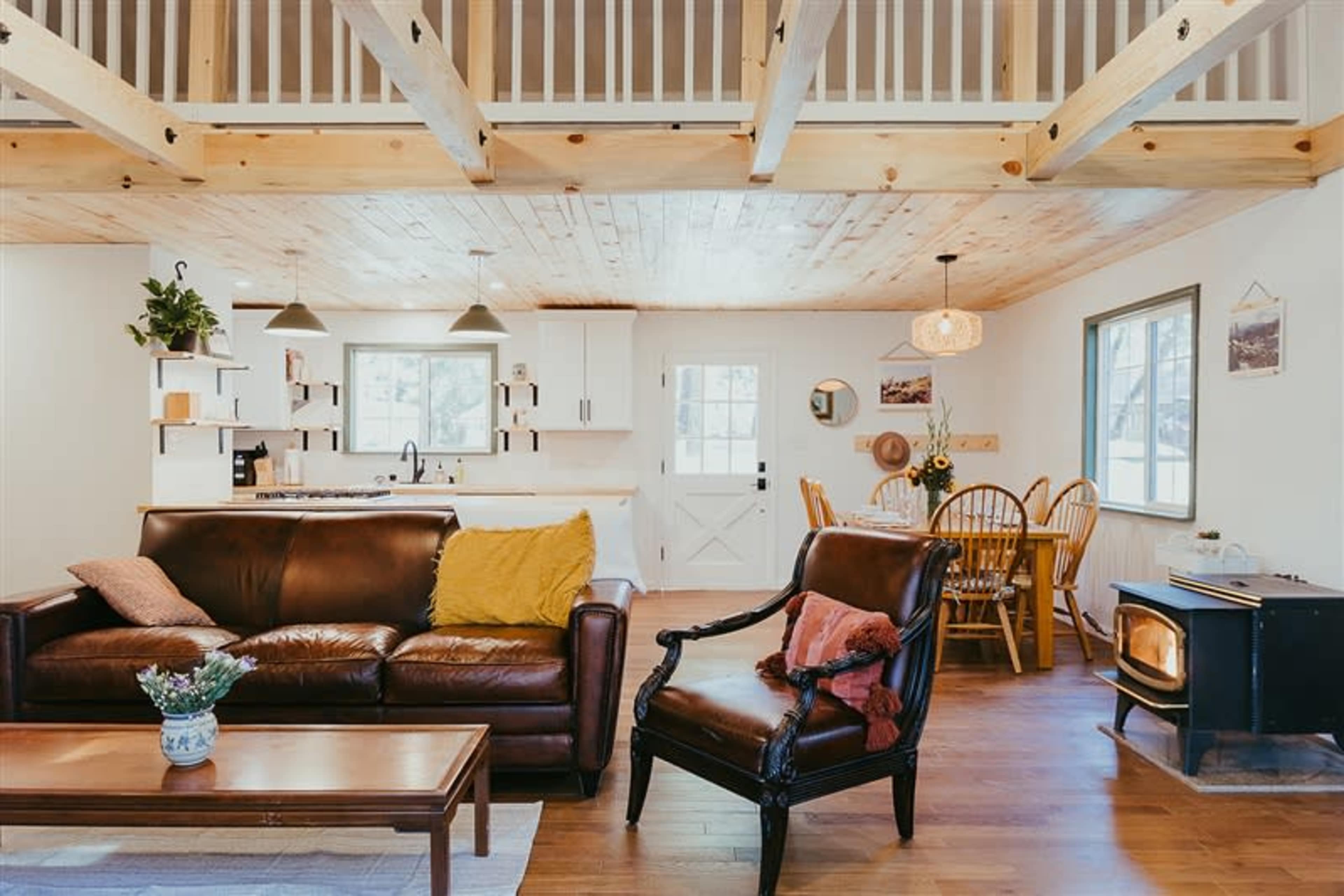 A cozy living space with a leather sofa, a dining area with wooden chairs, and a modern kitchen in a well-lit room featuring wooden beams and a wood stove.