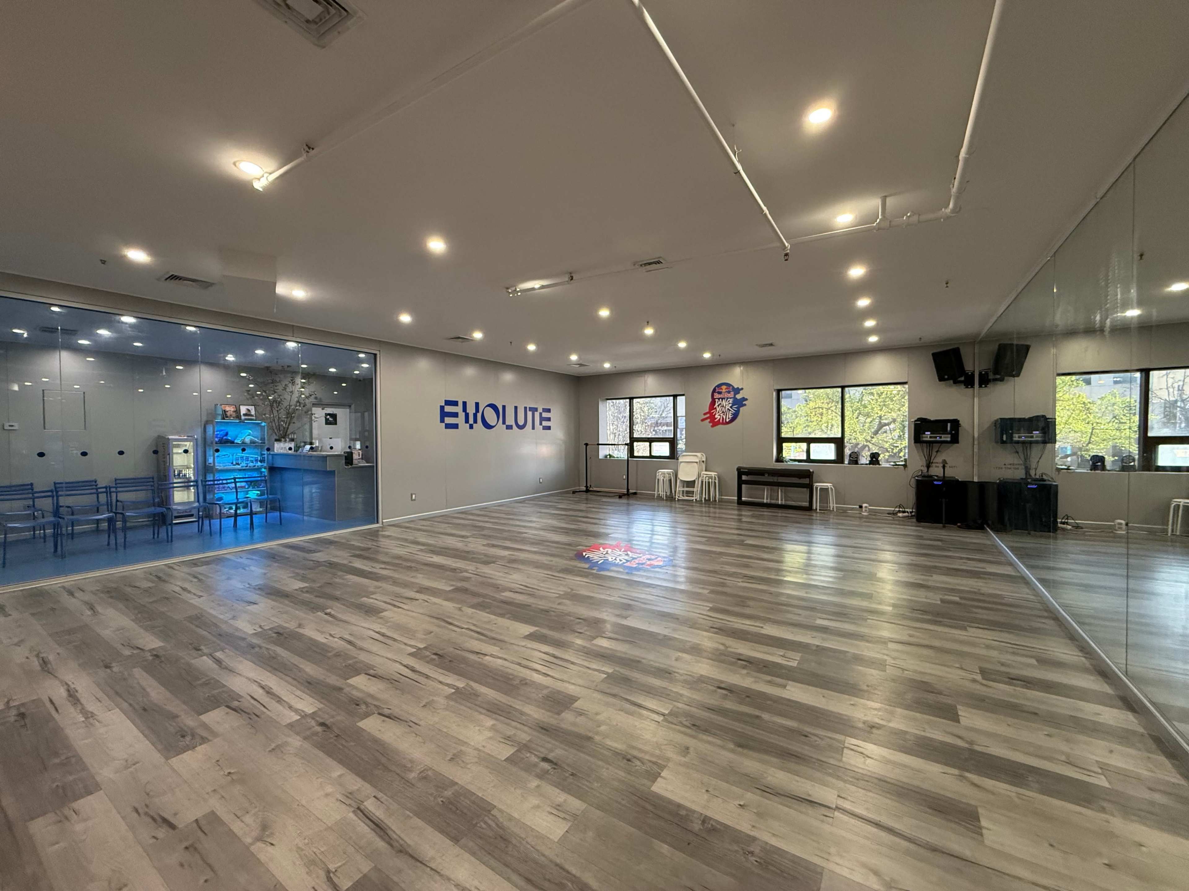 The image shows a spacious, well-lit studio with a wooden floor, featuring a modern design and a large glass wall displaying a room with blue furniture in the background.